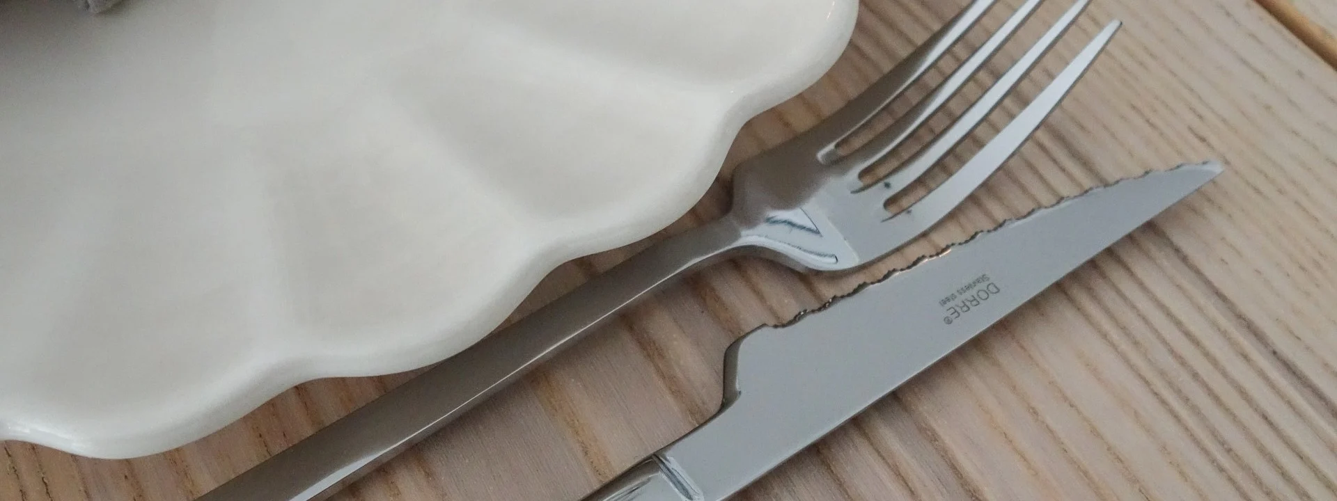 Elegant table setting with a white ruffled plate, grey linen napkin, gold napkin ring, silver fork and knife, and a wine glass on a wooden table.