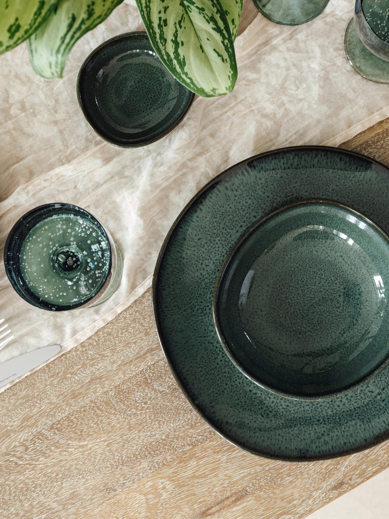 Table setting with dark green ceramic plates, bowls, and glasses on a light linen runner and wooden table, with a green plant.