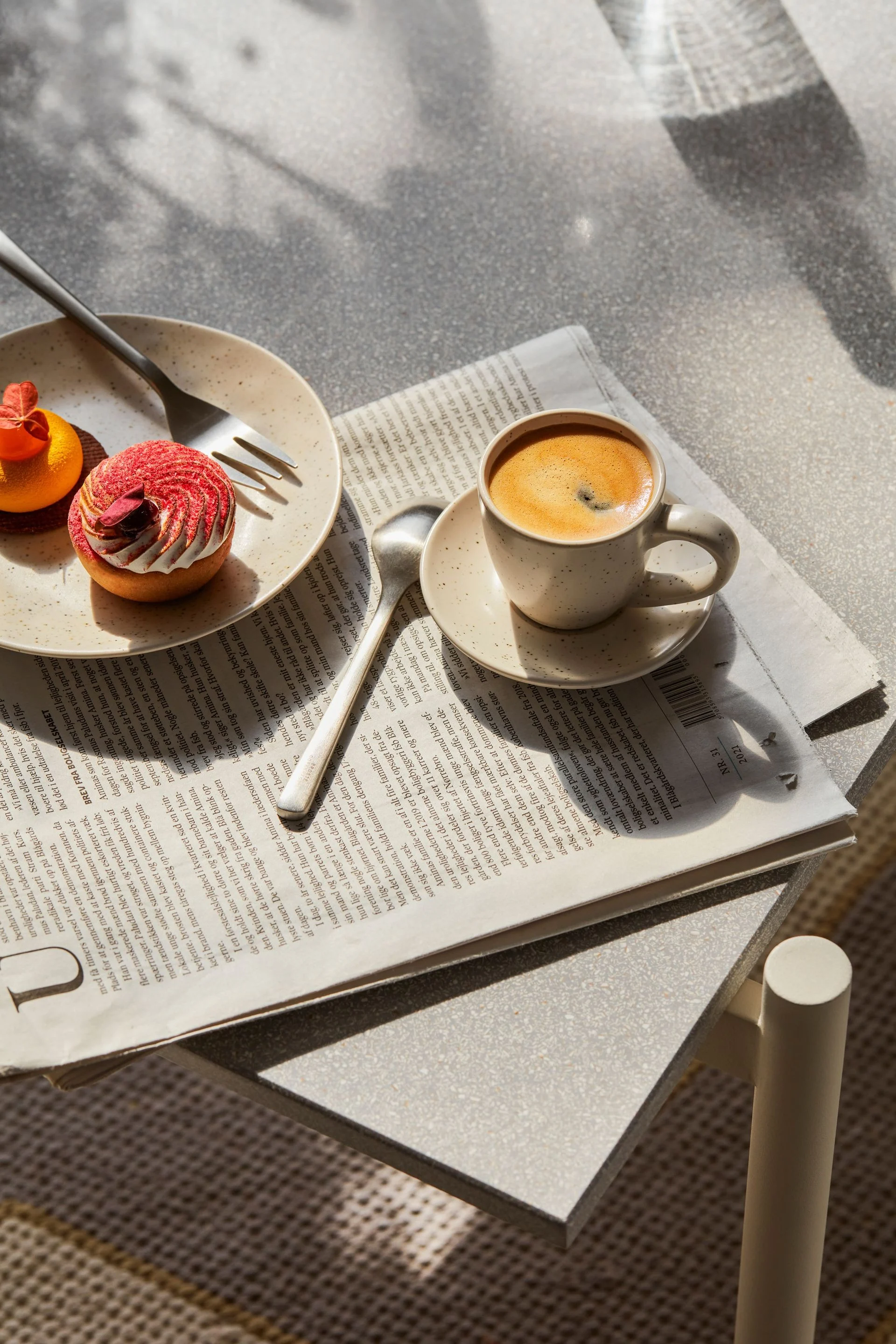 The Nordic Vanilla espresso cup from Broste Copenhagen on a breakfast table, with a daily newspaper underneath and a plate of cake next to it.
