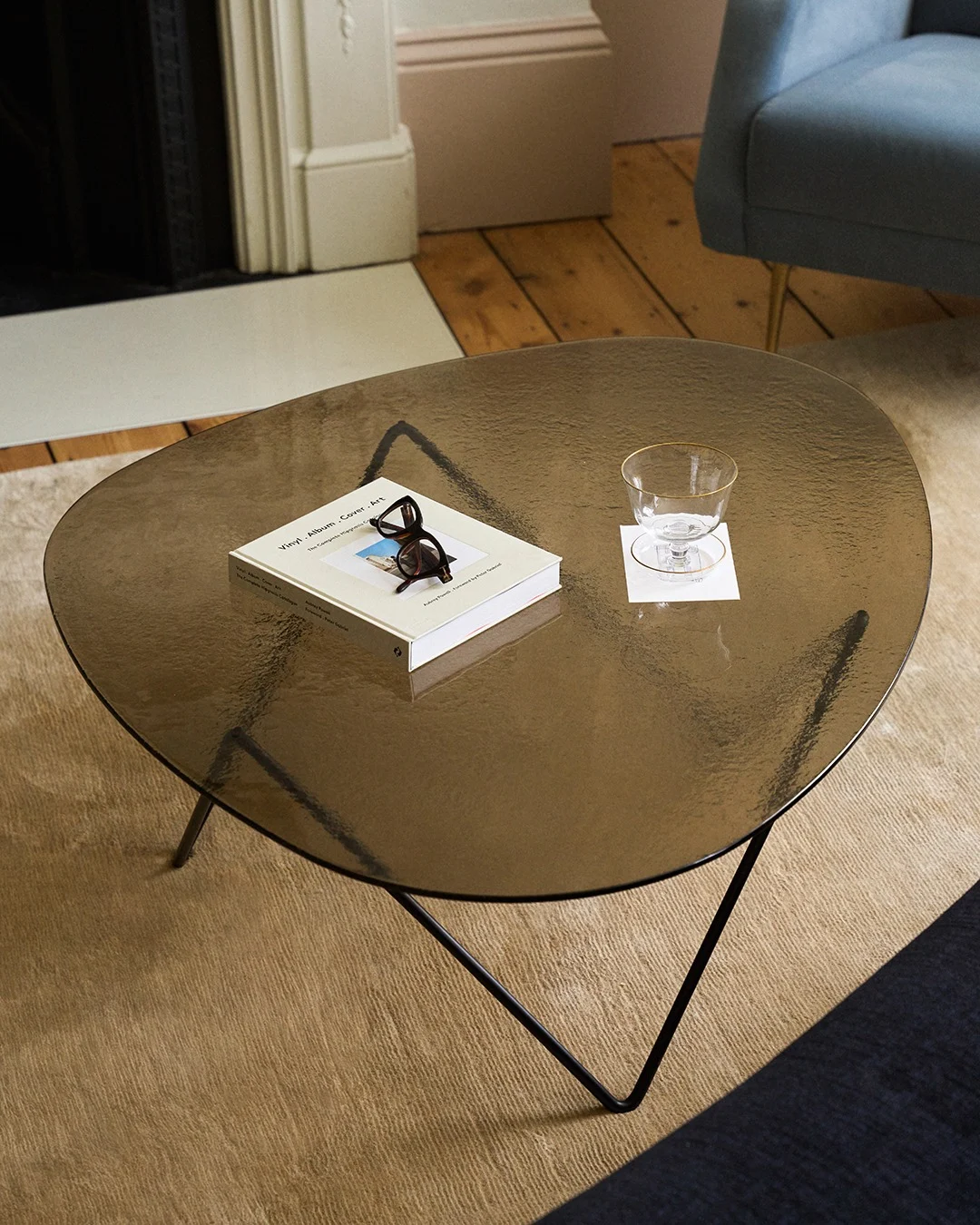 Organic-shaped dark glass coffee table with a book, tortoiseshell glasses, and a gold-rimmed glass on a coaster.