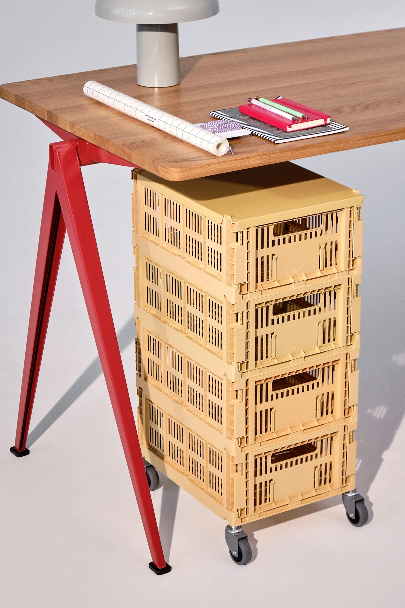 A wooden desk with red legs, a lamp, paper, notebooks, and pens on top, and a stack of four yellow storage crates on wheels underneath.