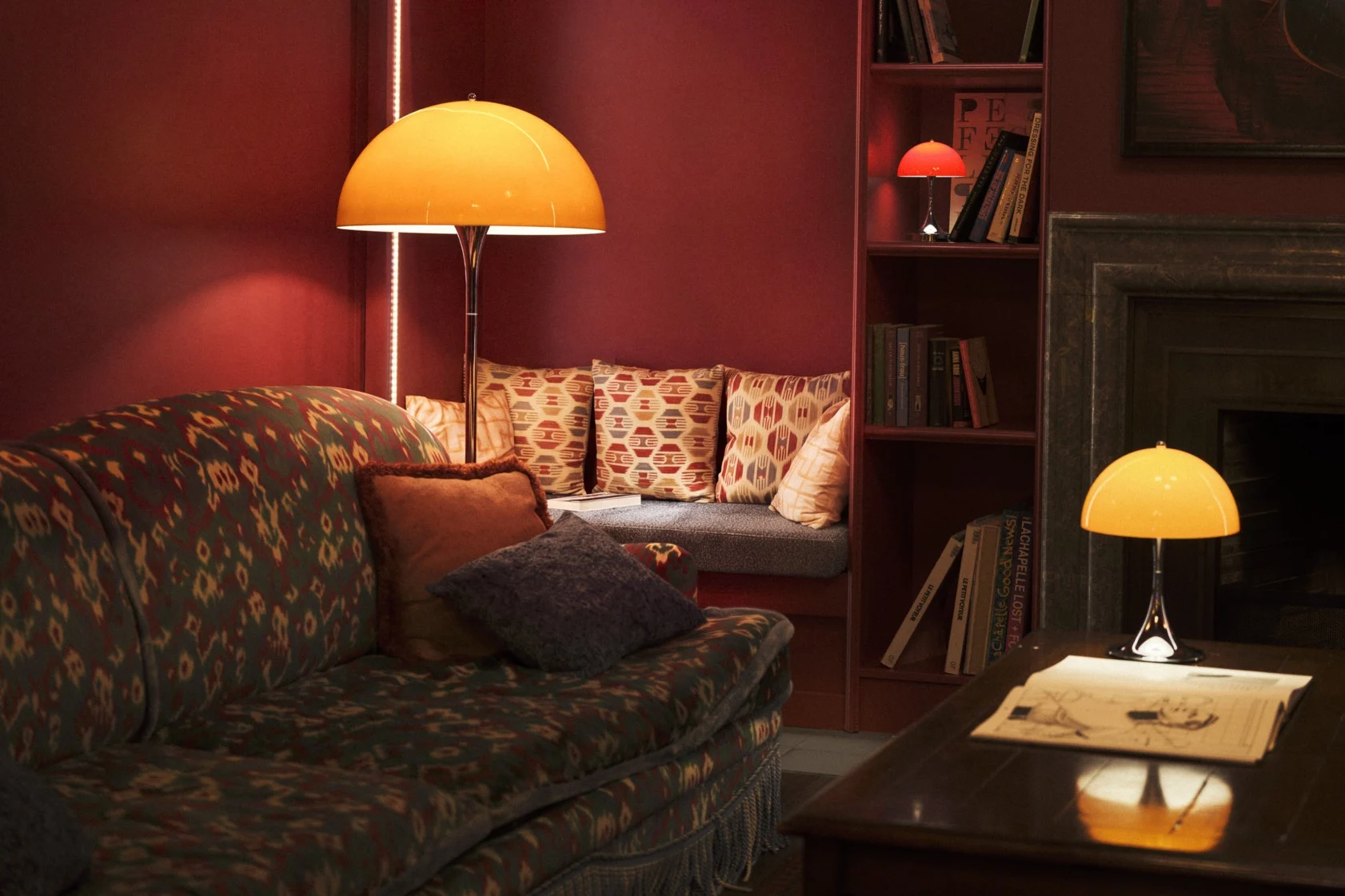 A retro floor lamp with an orange dome shade and chrome stand illuminates a room with a patterned rug and wooden wardrobe.