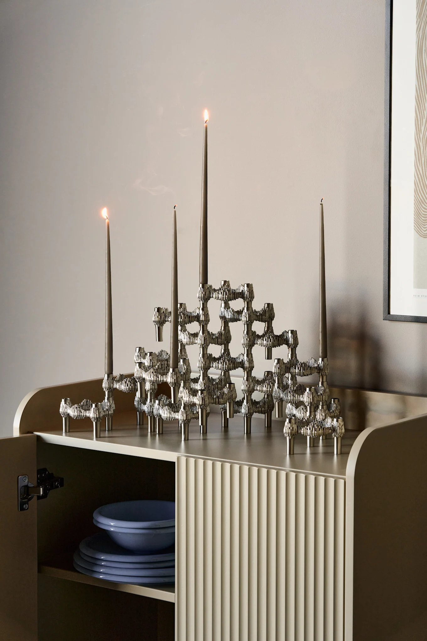 Modular silver candle holder with grey taper candles on a beige cabinet. Blue plates are visible inside the open cabinet.