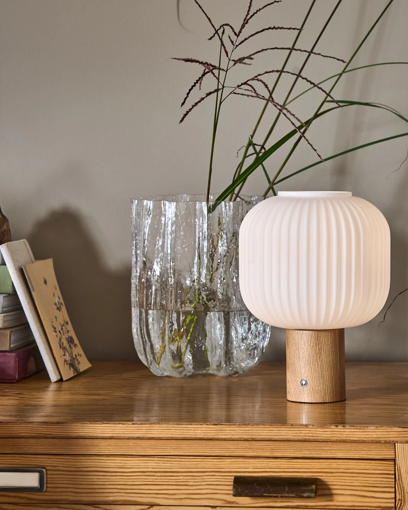 A ribbed, mushroom-shaped lamp with a wooden base next to a textured glass vase with plants and a stack of books on a wooden dresser.