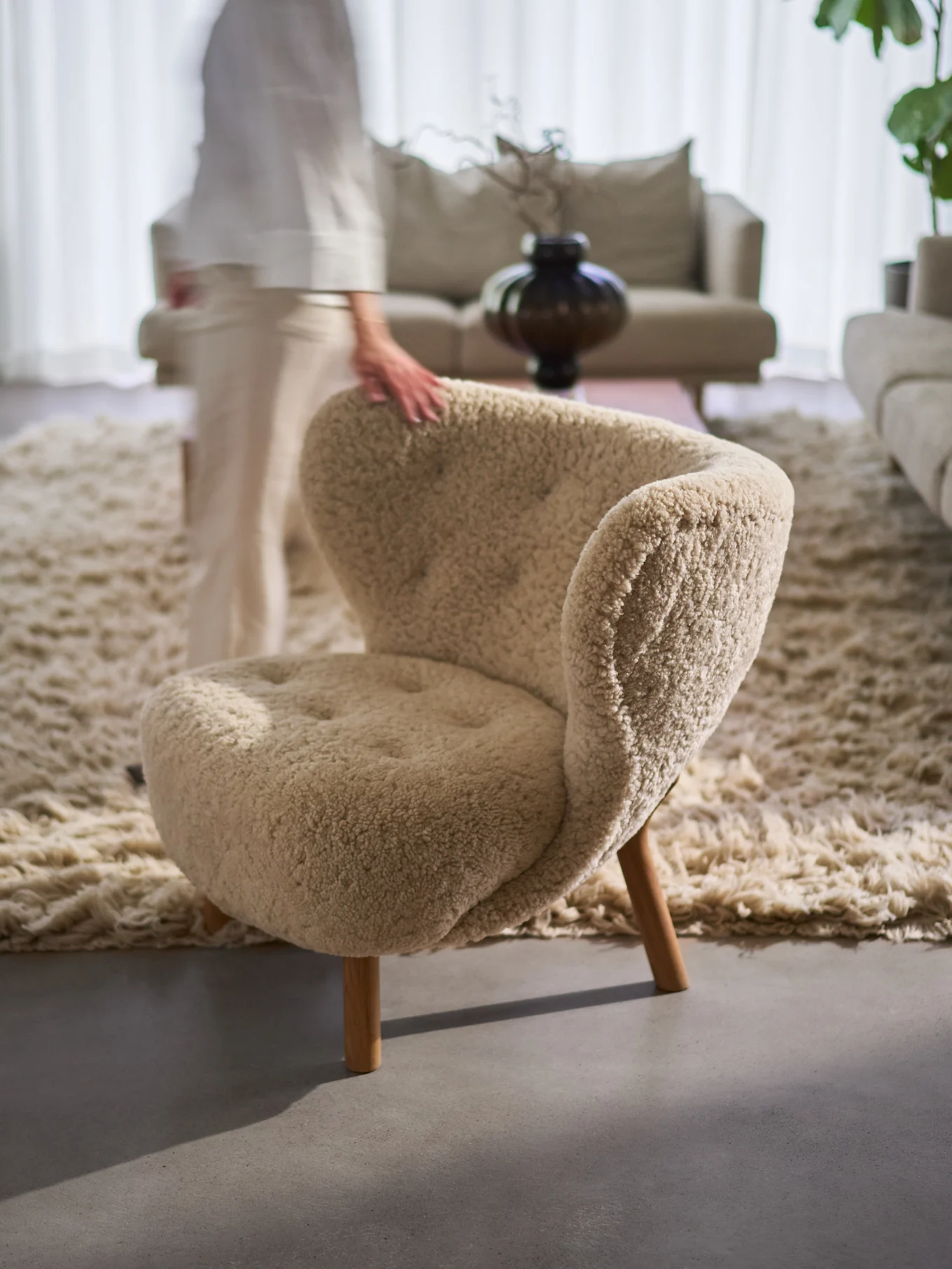 A person's hand rests on a cozy, textured beige armchair with wooden legs, on a shaggy rug in a living room.