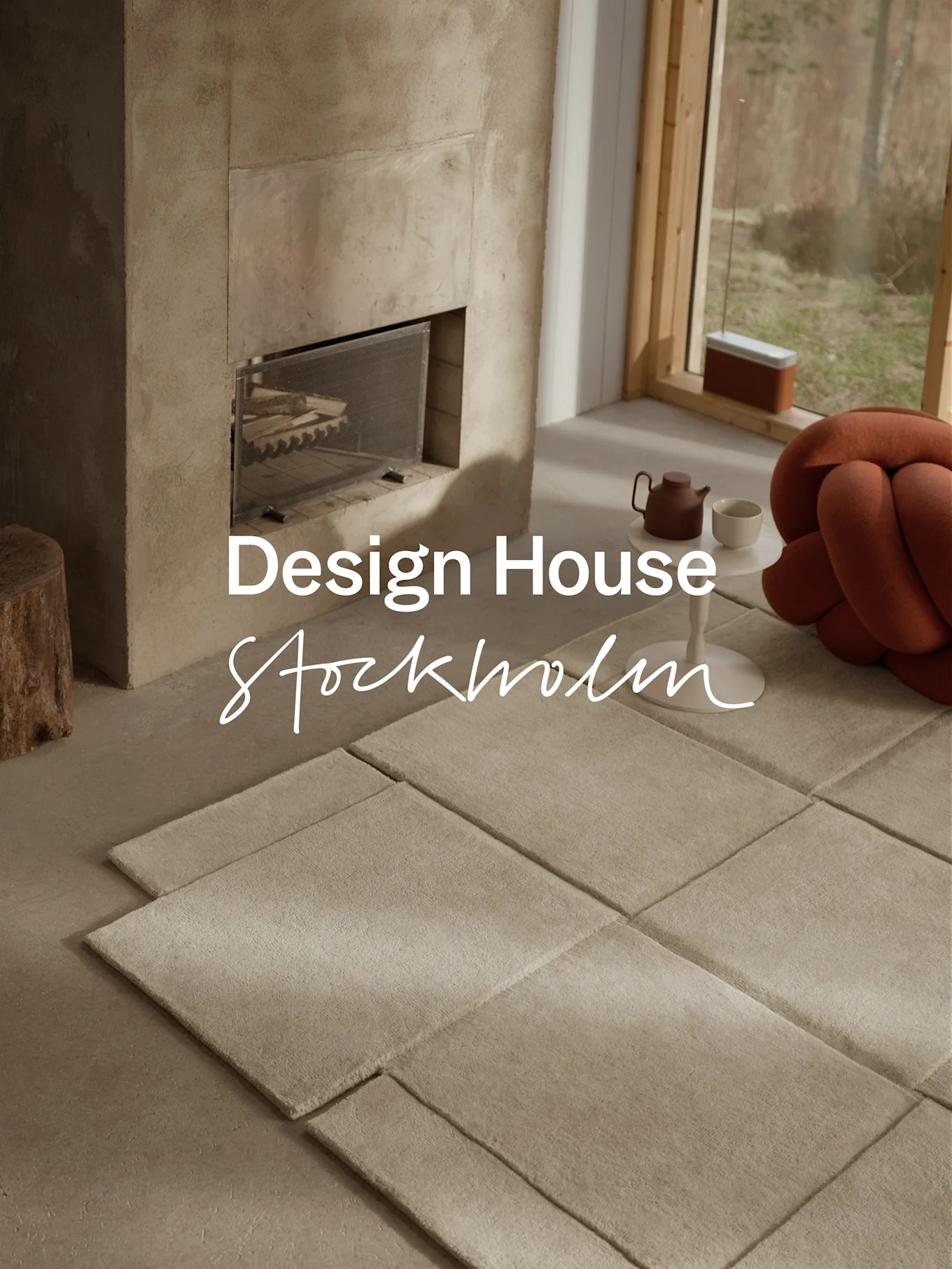 Modern living room with concrete fireplace, modular light-colored rugs, a teapot, and an orange knotted cushion next to a large window.