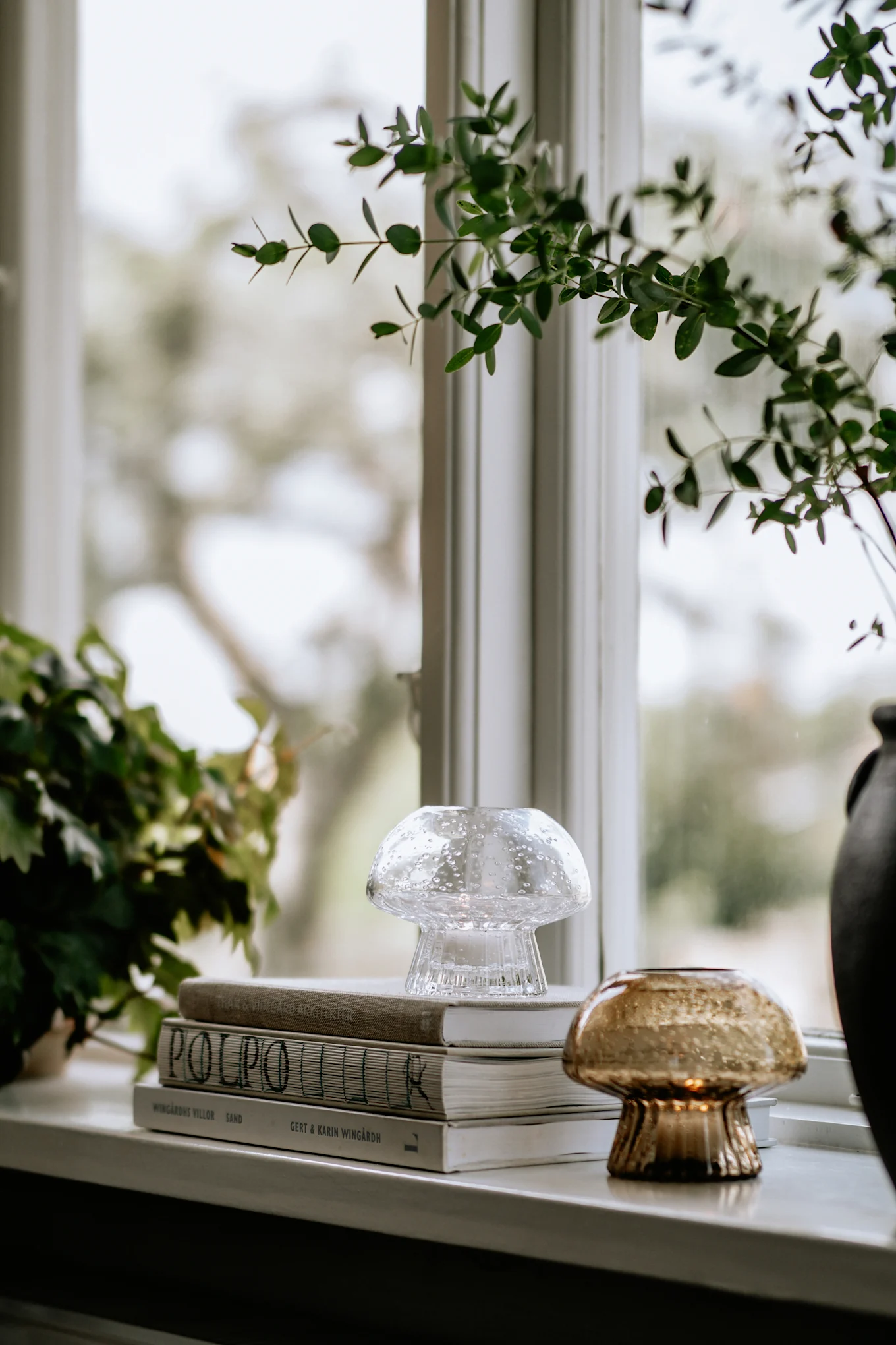 Clear bubble glass and amber glass tealight holders, books, and green plants on a windowsill.
