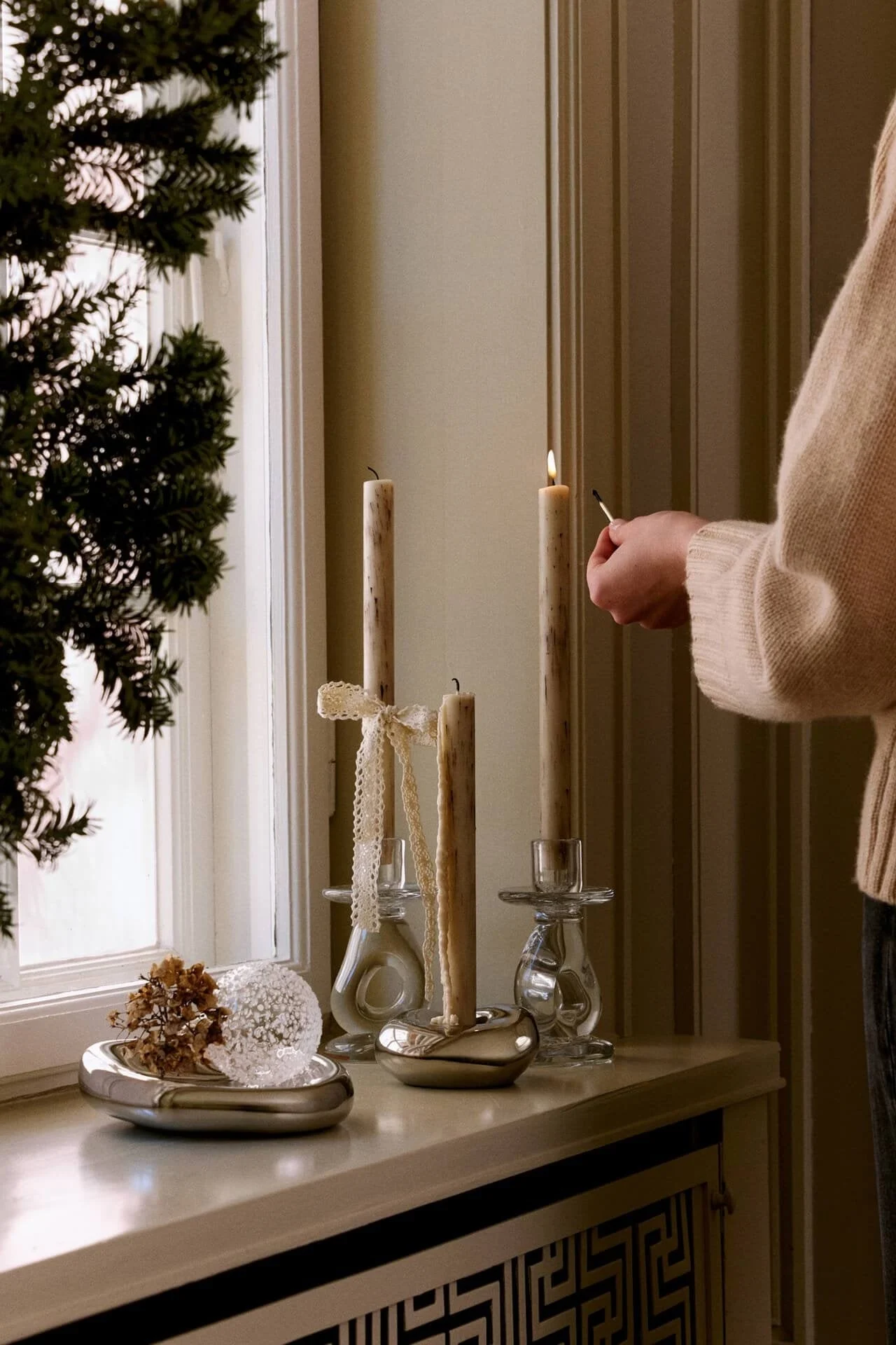 A person lights a tall, textured candle on a windowsill with other candles, a silver dish of dried flowers, and a clear sphere.