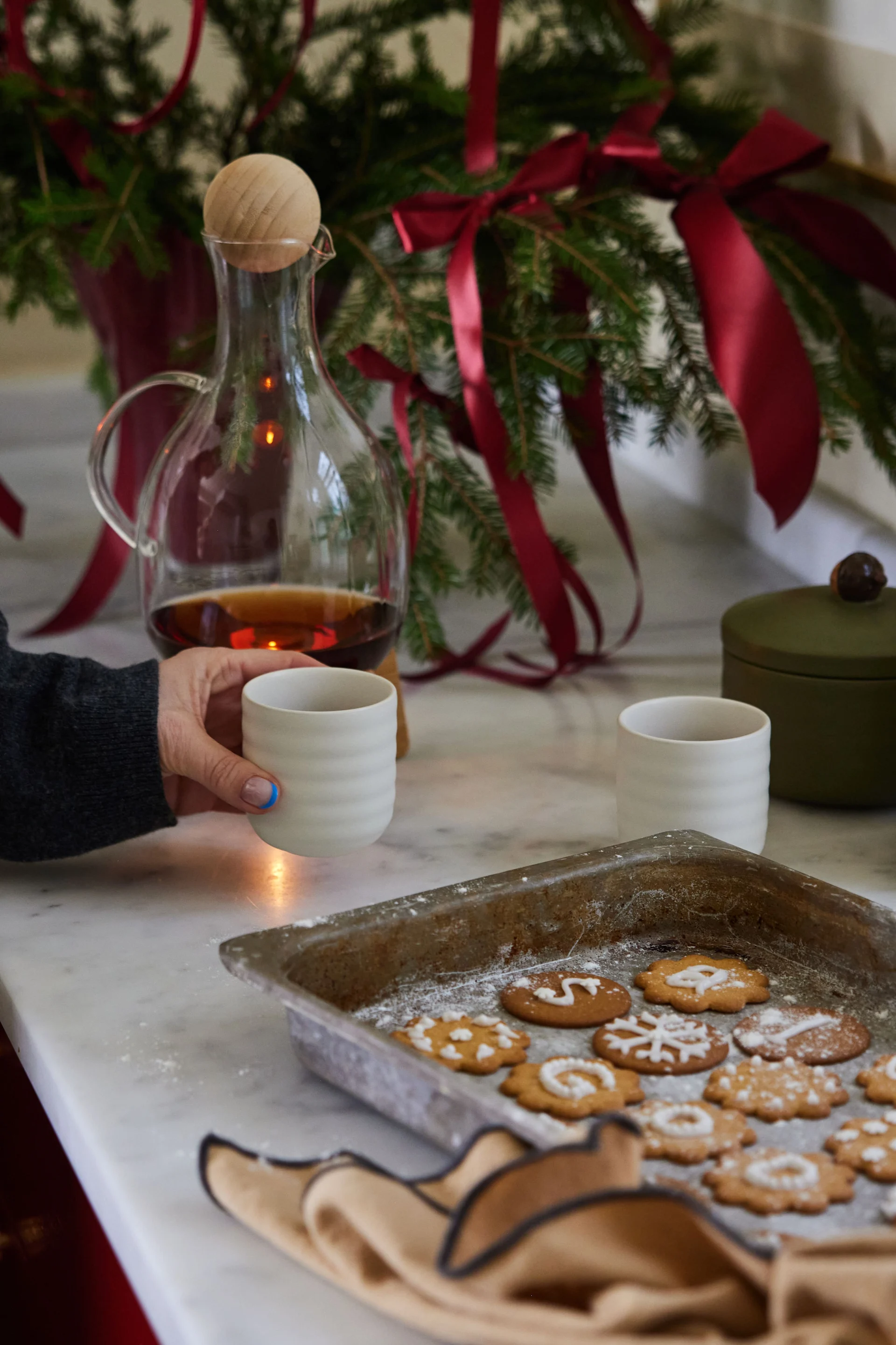A hand holds a mug of glögg next to a tray of pepparkaka, one of many Swedish Christmas traditions. 