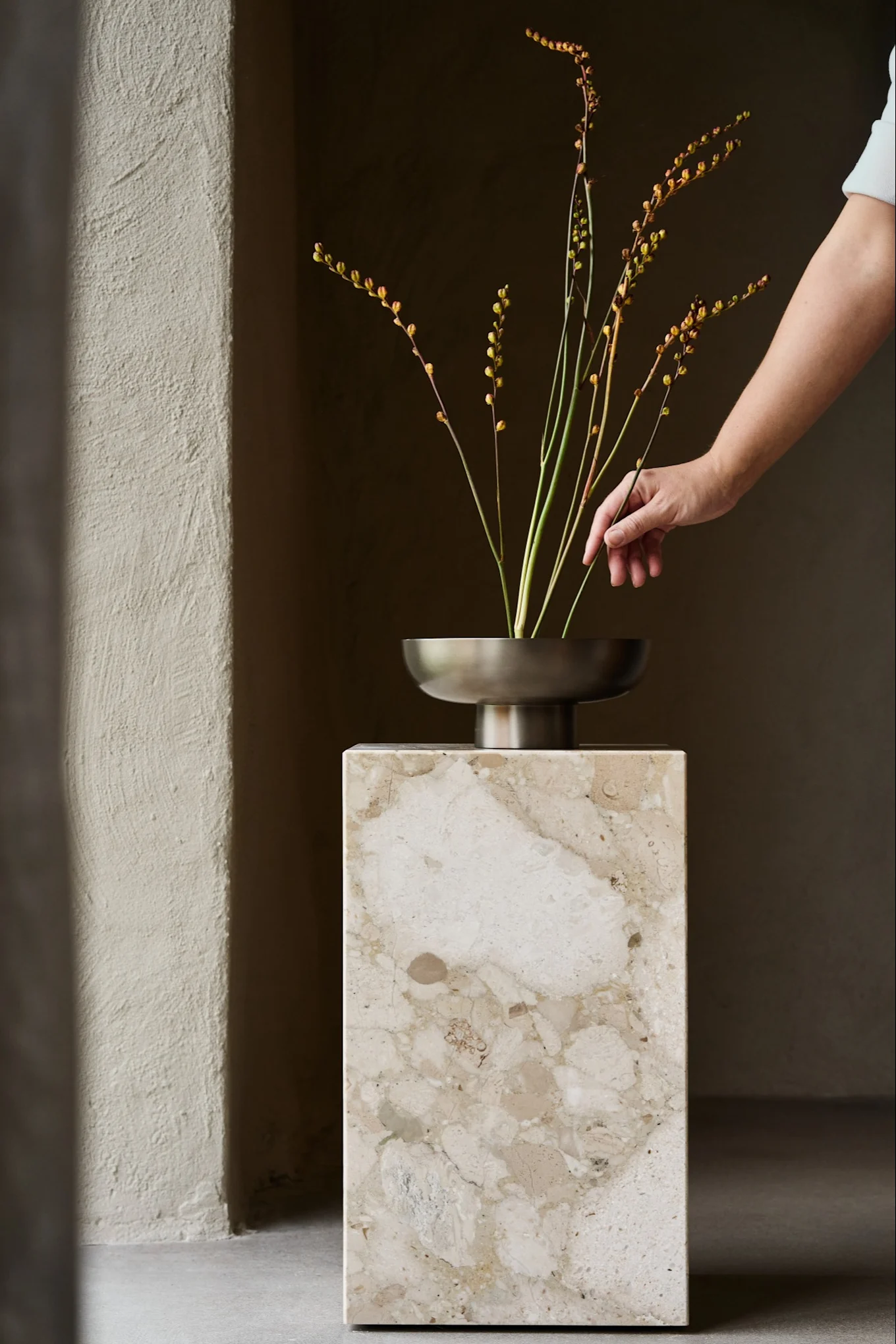 Hand arranging delicate yellow-budded stems in a bronze bowl on a beige marble pedestal.