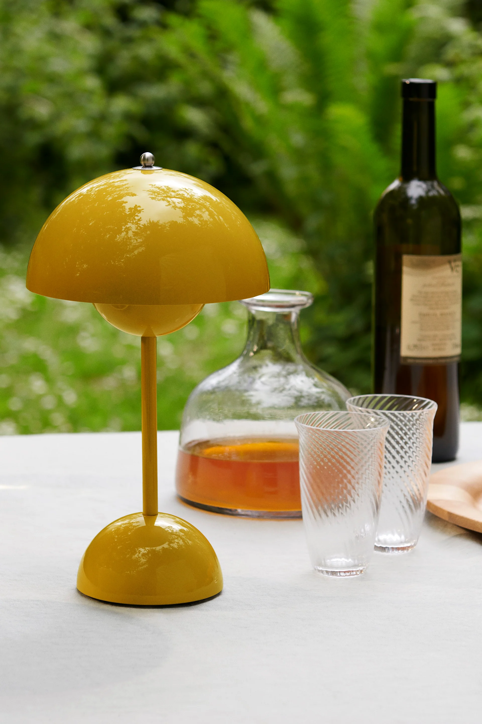 A bright yellow, mushroom-shaped table lamp sits on a white table, with a carafe, two patterned glasses, and a wine bottle behind it, against a green background.