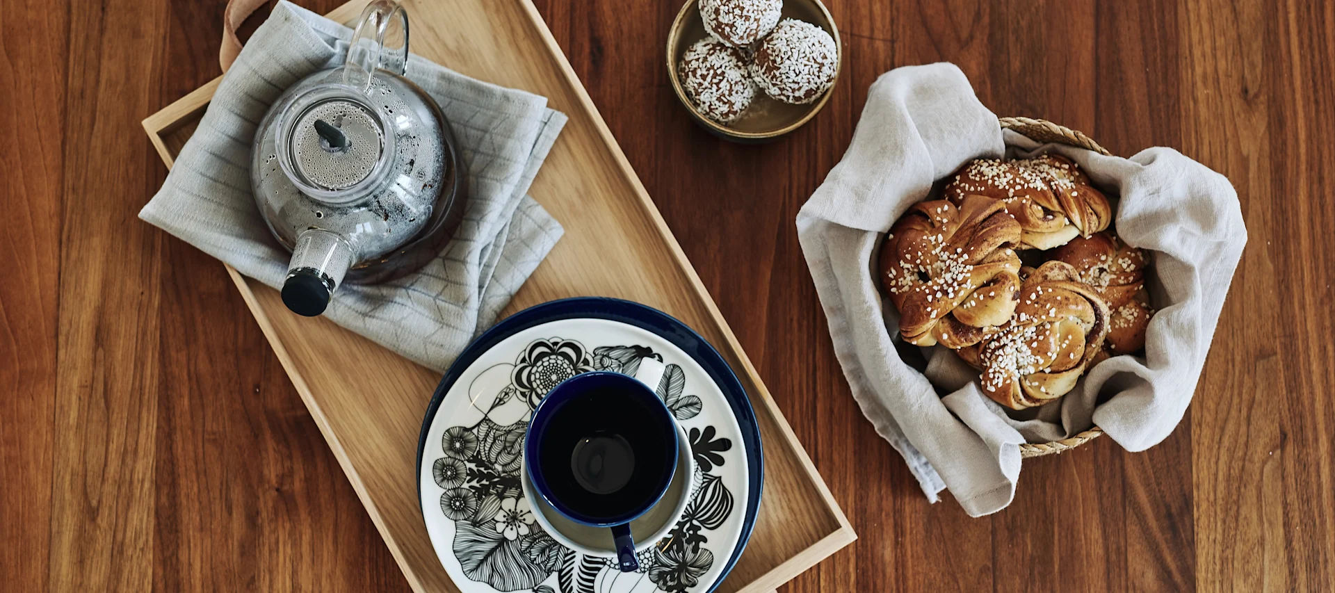 Swedish fika - here you see a glass teapot on a tea towel from Swedish Grace with a selection of mugs and plates and cinnamon buns.