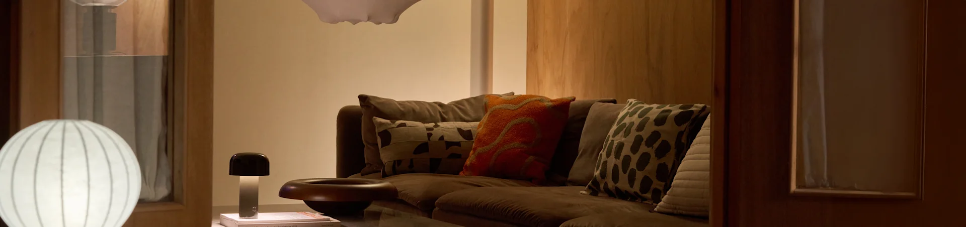 A cozy living room with a brown sofa, various patterned pillows, and two lamps: a large white ribbed floor lamp and a small mushroom-shaped table lamp.