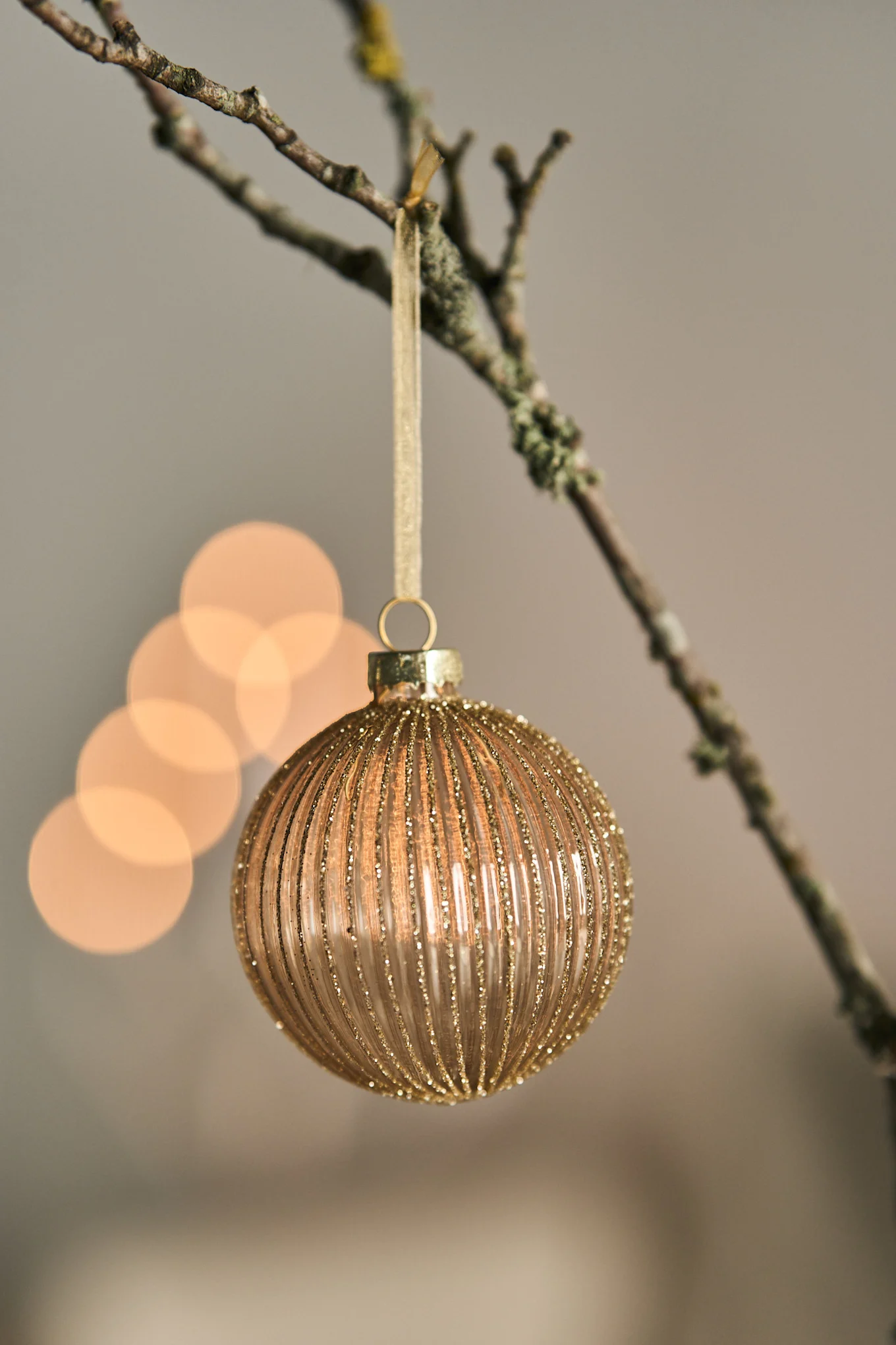 A minimalist Christmas bauble hanging on a branch with twinkling lights behind. 