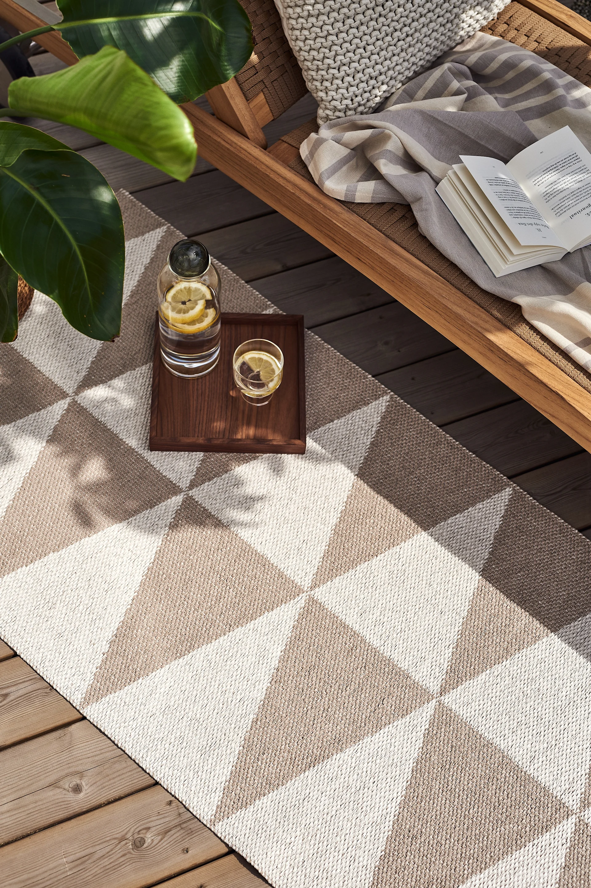 Overhead view of an outdoor lounge area with a geometric patterned rug, a wooden lounge chair with a book and blanket, a plant, and a tray with lemon water.