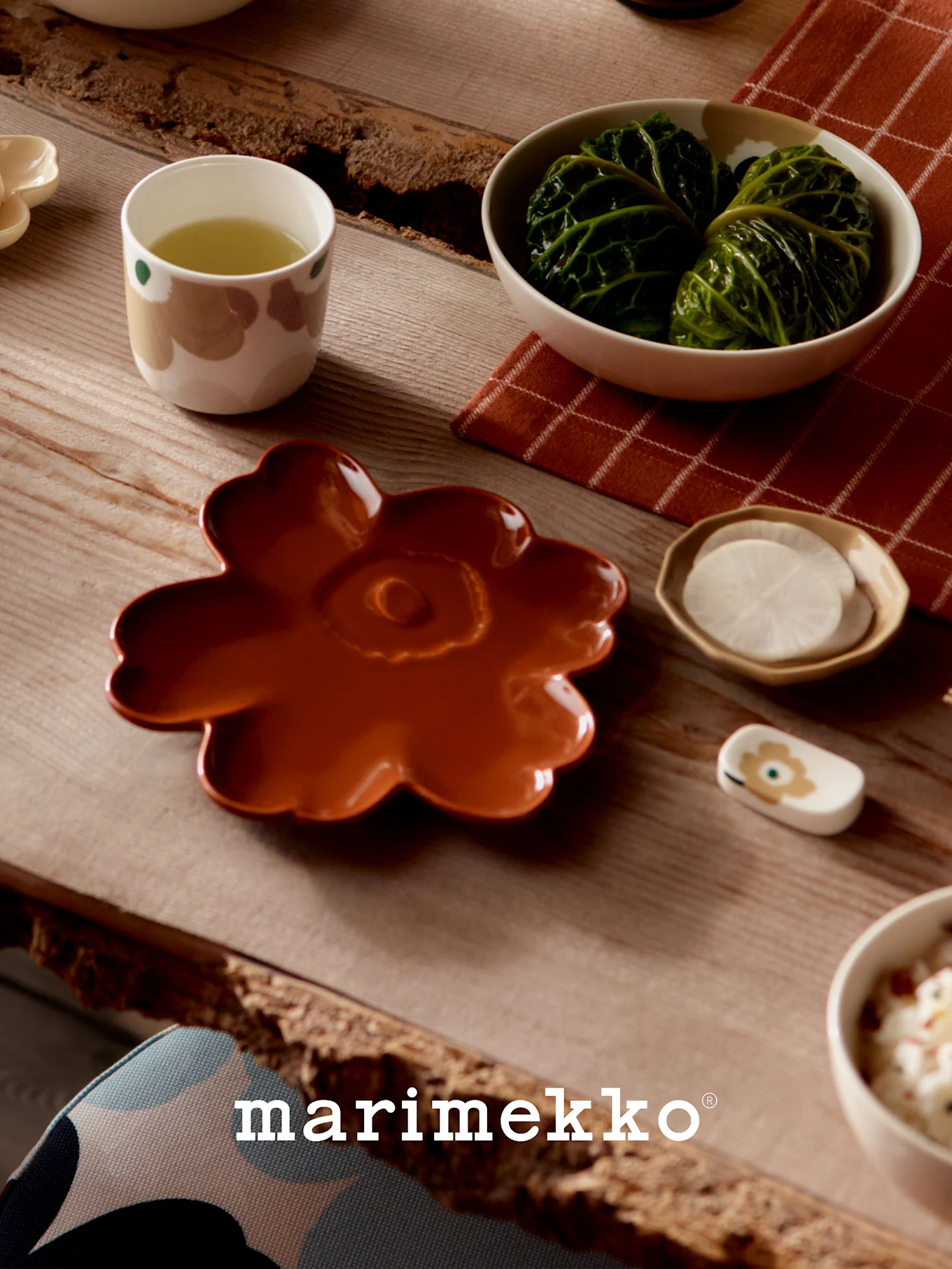Table set with Marimekko dishes: a rust-colored flower plate, a cup of green tea, cabbage rolls, and sliced radishes.