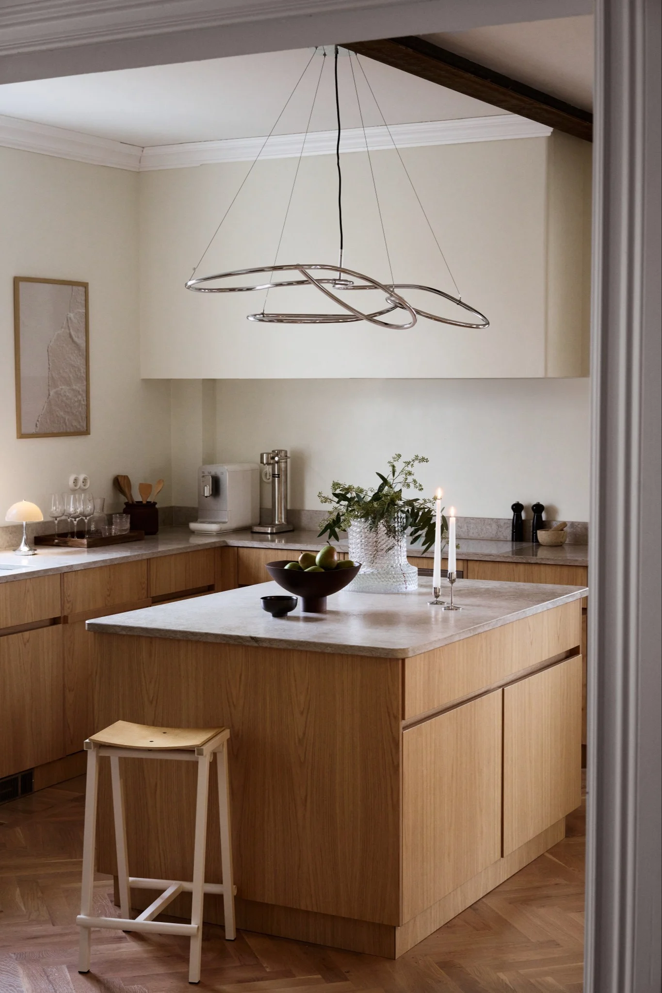 Modern kitchen with wood cabinets, light countertops, and a unique chrome ring chandelier over an island with fruit and candles.