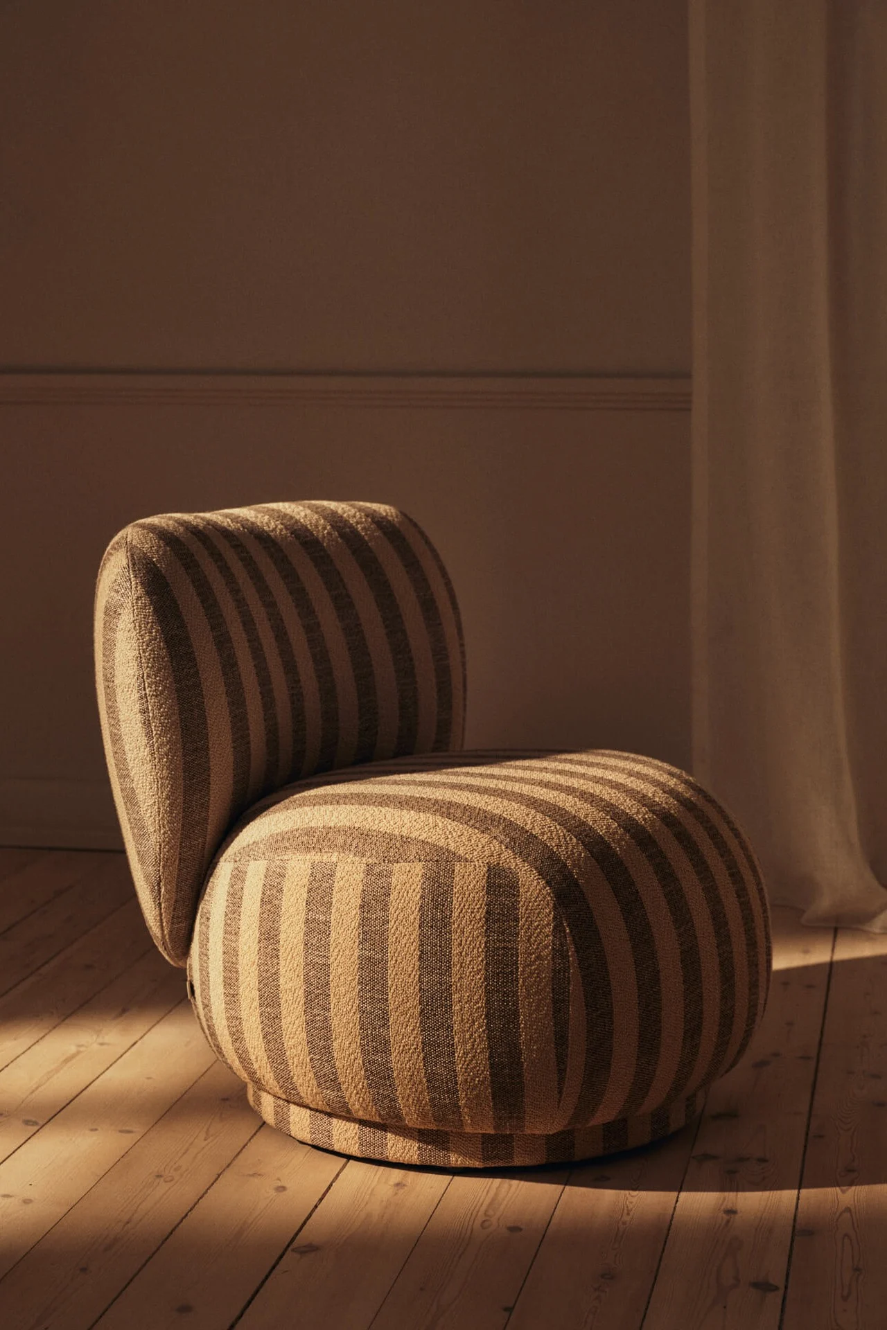Modern, rounded armchair with beige and brown stripes on a wooden floor, bathed in warm light.