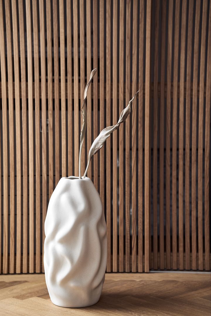 Dark woods and straight lines that meet round shapes are some of the autumn interior design trends 2021. Here the Drift vase from Cooee Design in beige stands in front of a dark wood panel.