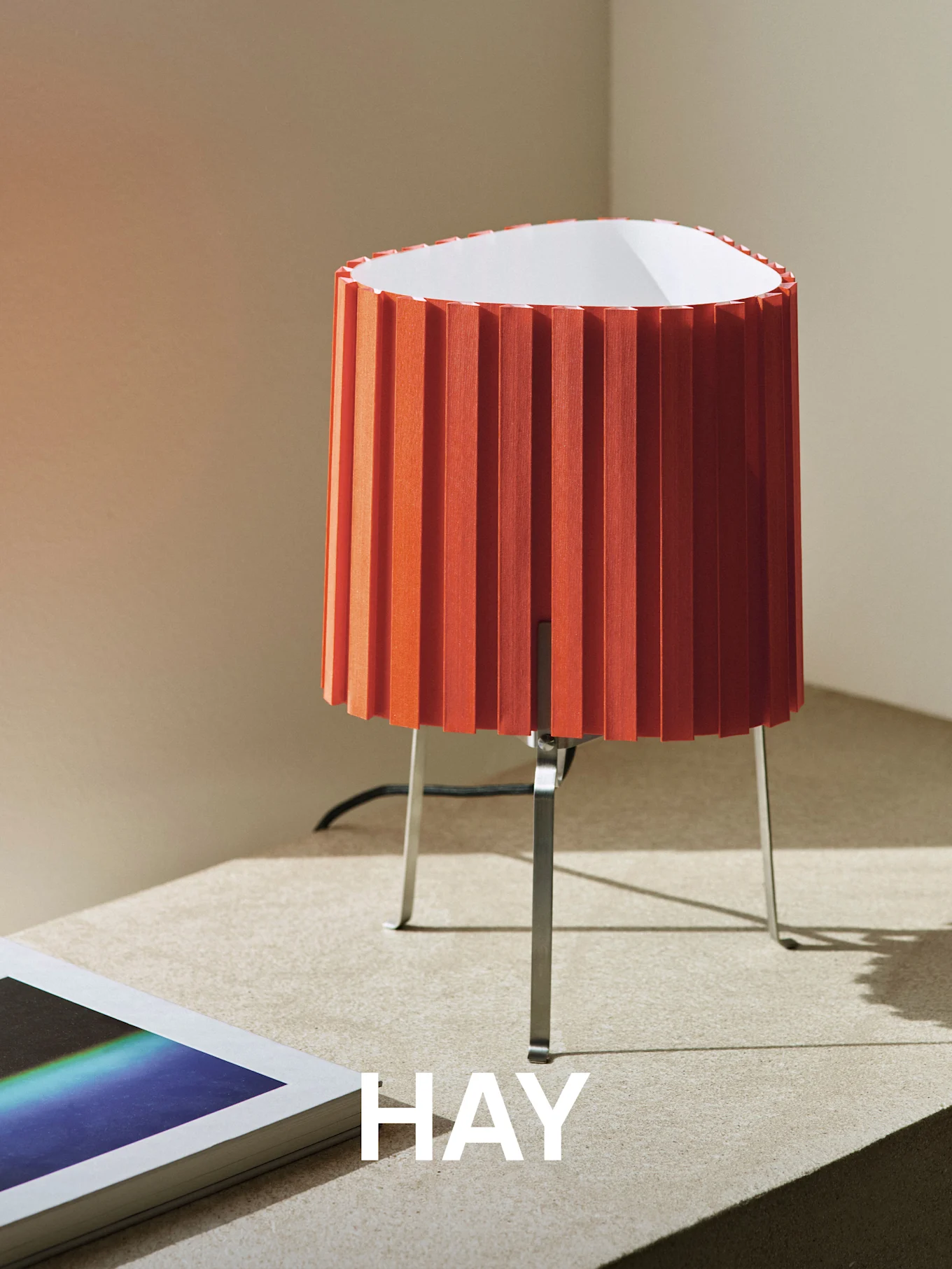 Orange pleated table lamp with a metal base on a textured surface, next to a book. The word 'HAY' is overlaid.