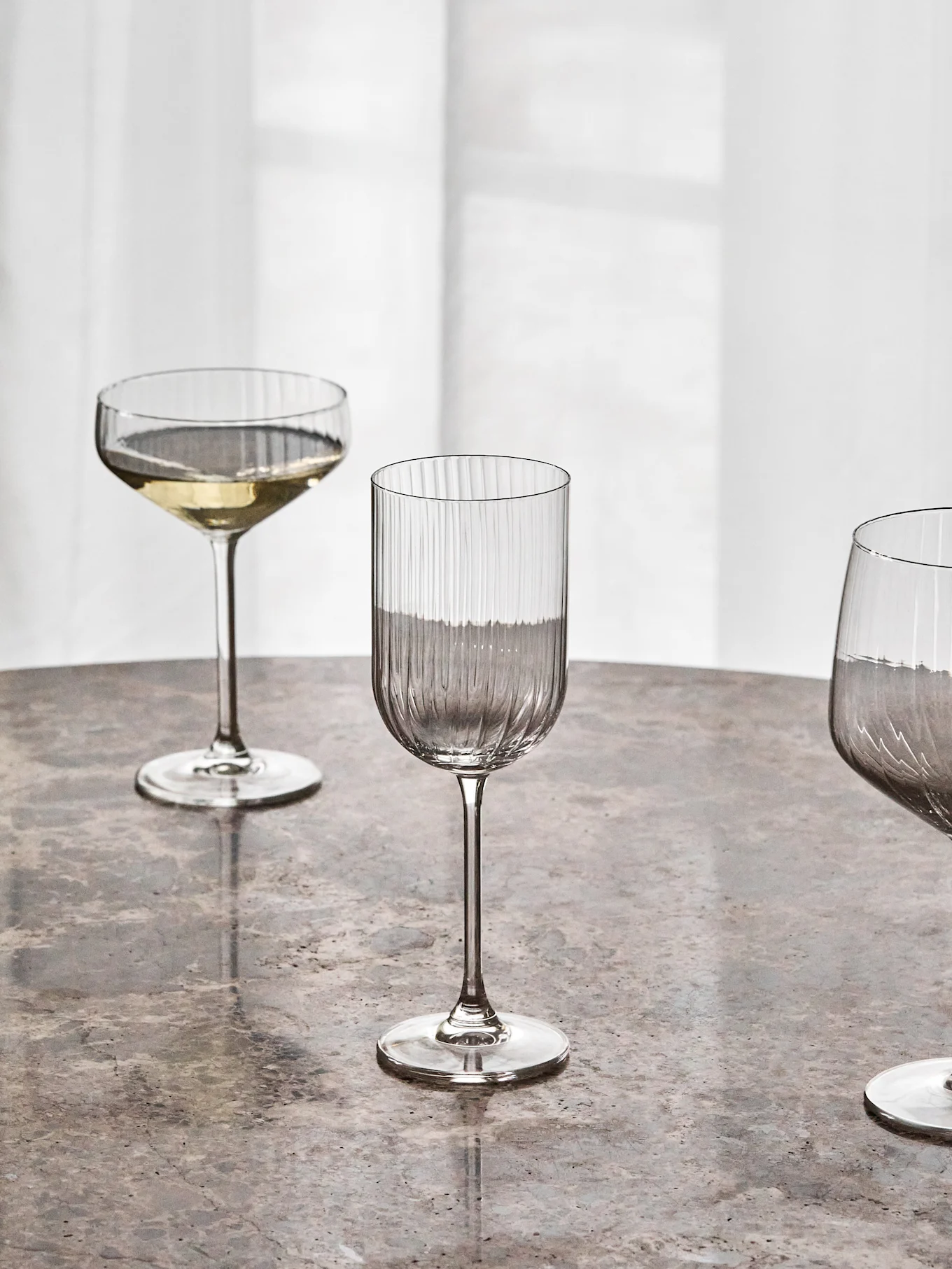 Three clear stemmed glasses on a marble table, including a coupe with white wine and a tall ribbed glass.