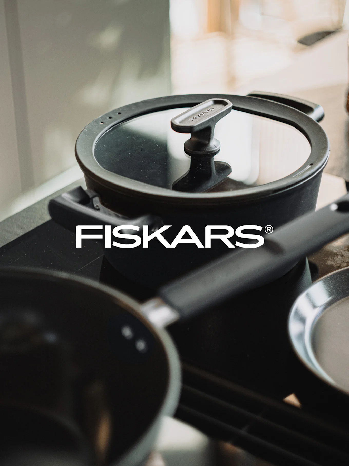 A black cooking pot with a glass lid and the white Fiskars logo on a stove, with a blurred frying pan in the foreground.