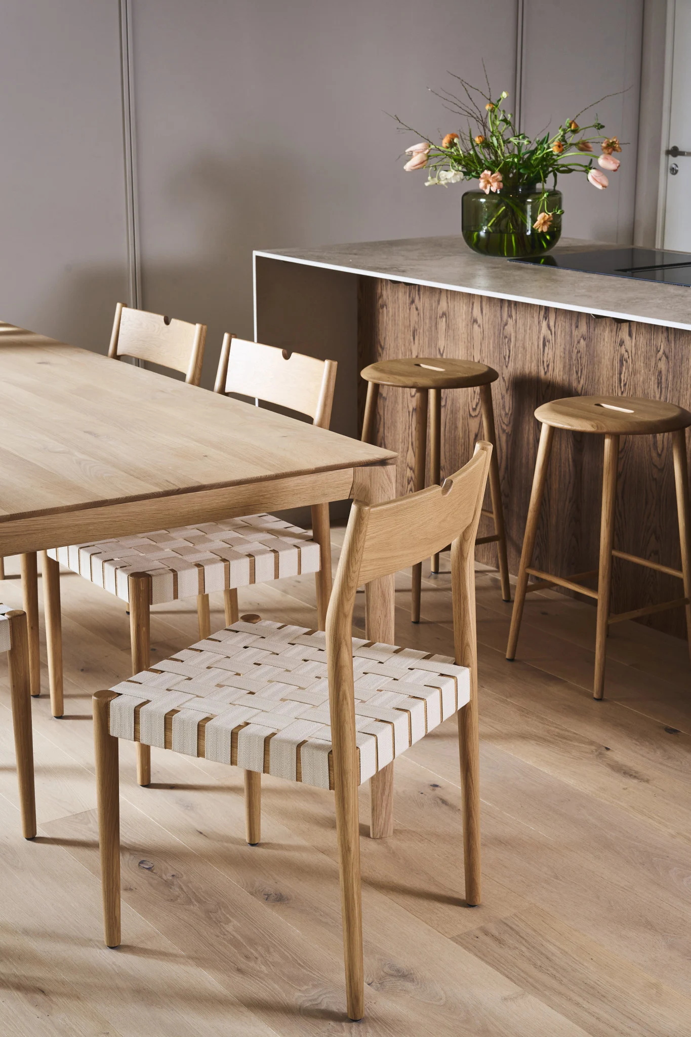 A modern dining room and kitchen with a light wooden table, chairs with woven seats, a wood-paneled island, and bar stools.