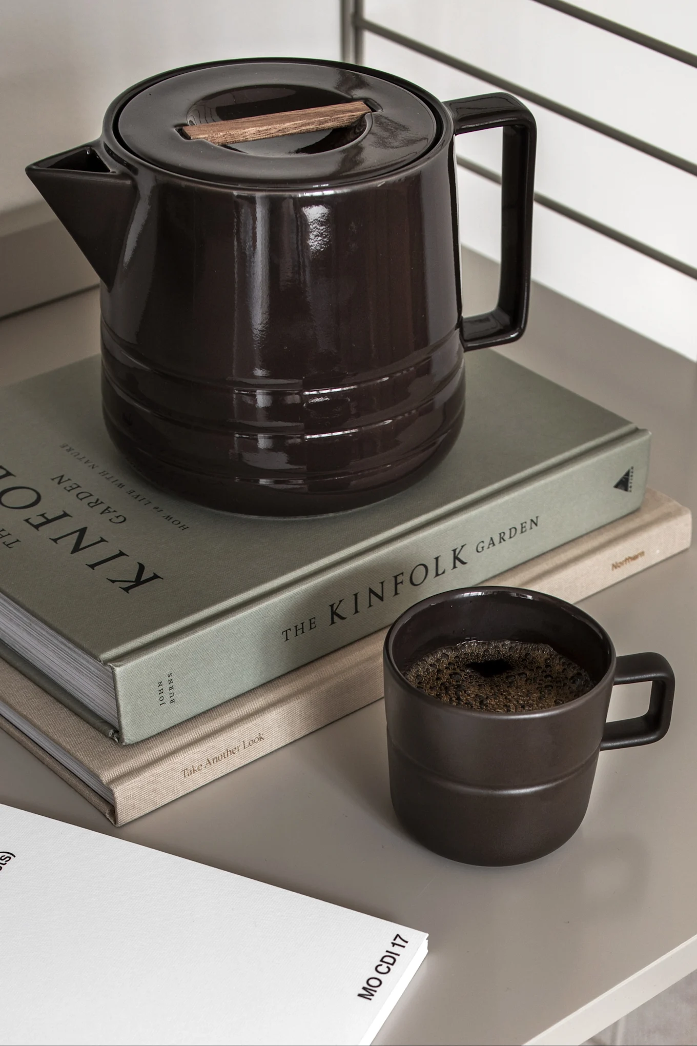 Dark brown ceramic coffee pot with wooden lid accent, a matching mug with coffee, and two books on a light surface.