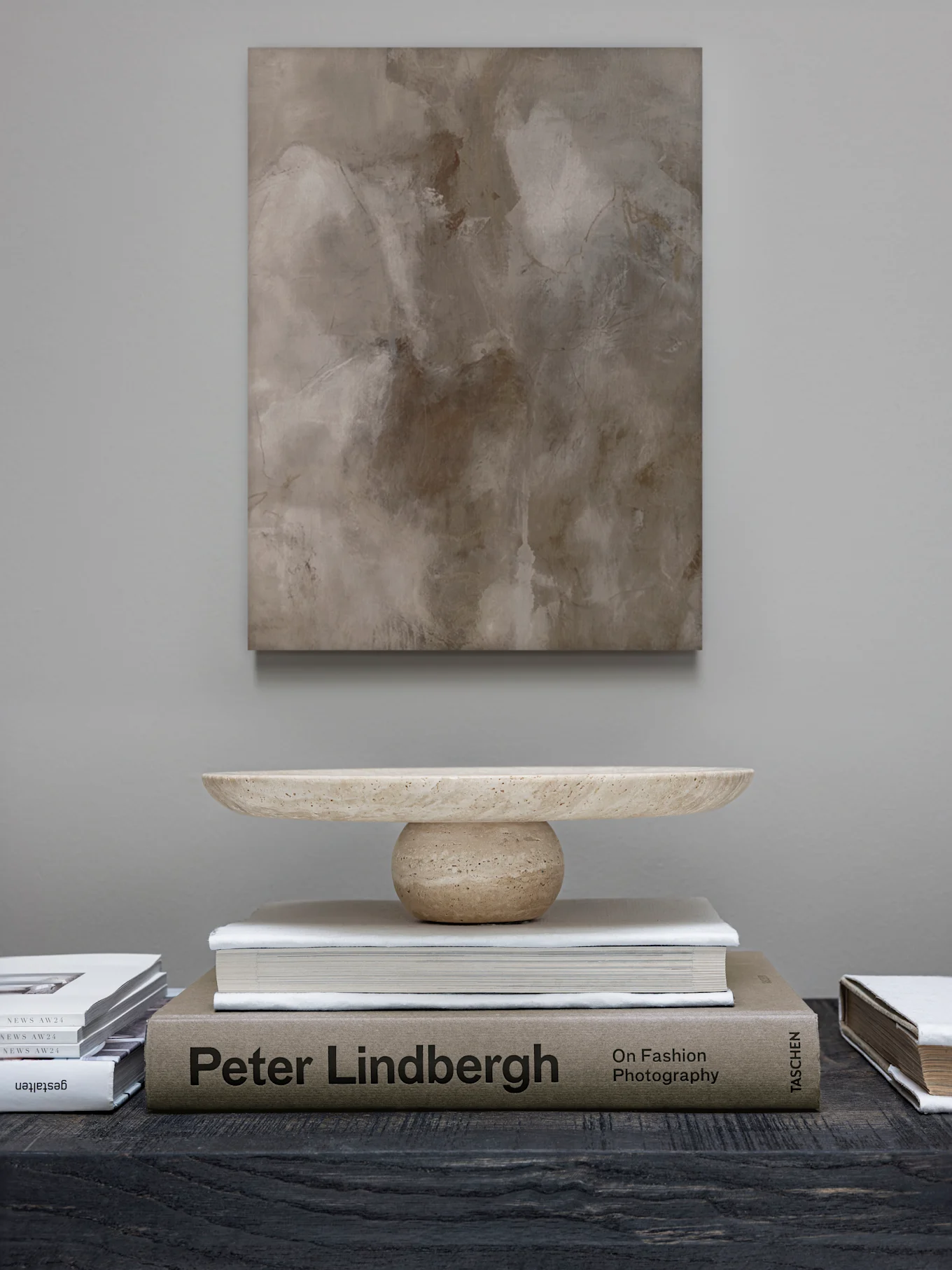 An abstract painting hangs above a light stone decorative bowl on a stack of books, including 'Peter Lindbergh,' on a dark table.