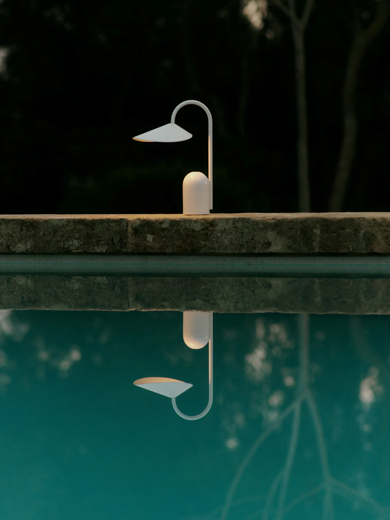 White modern lamp on a pool edge, reflected in blue water against a dark, blurred natural background.