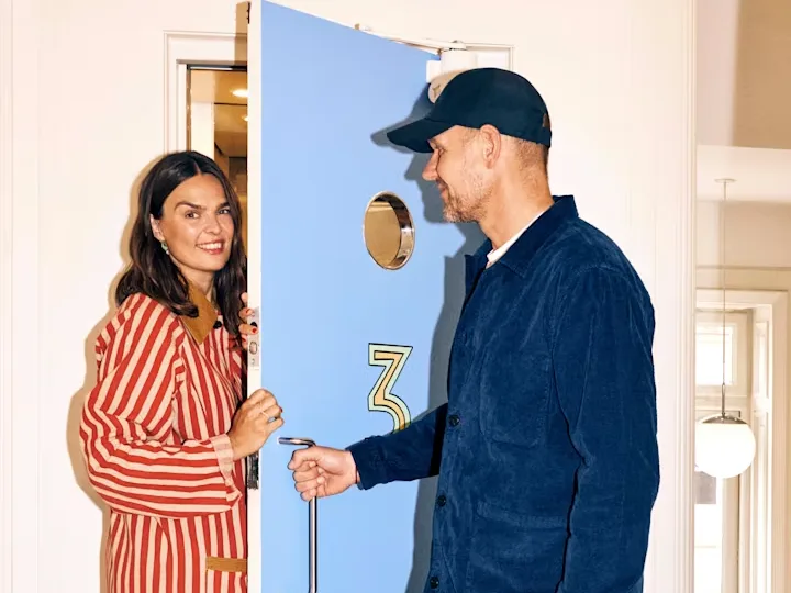 A smiling woman in a striped top peeking from behind a light blue door as a man in a cap opens it.