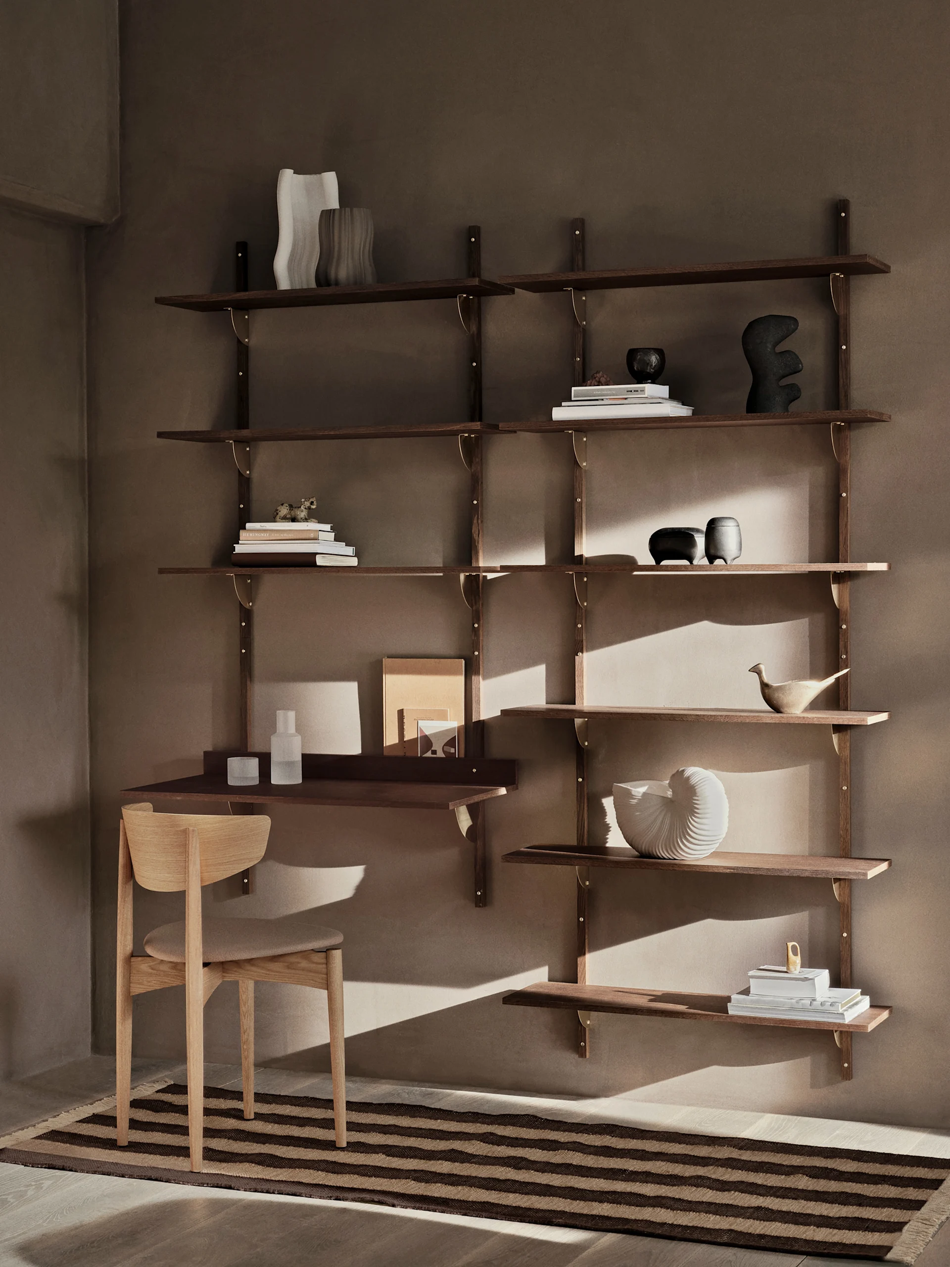 A modern dark wood wall shelving unit with a desk and chair, adorned with books and decorative objects, sits against a warm brown wall with a striped rug.