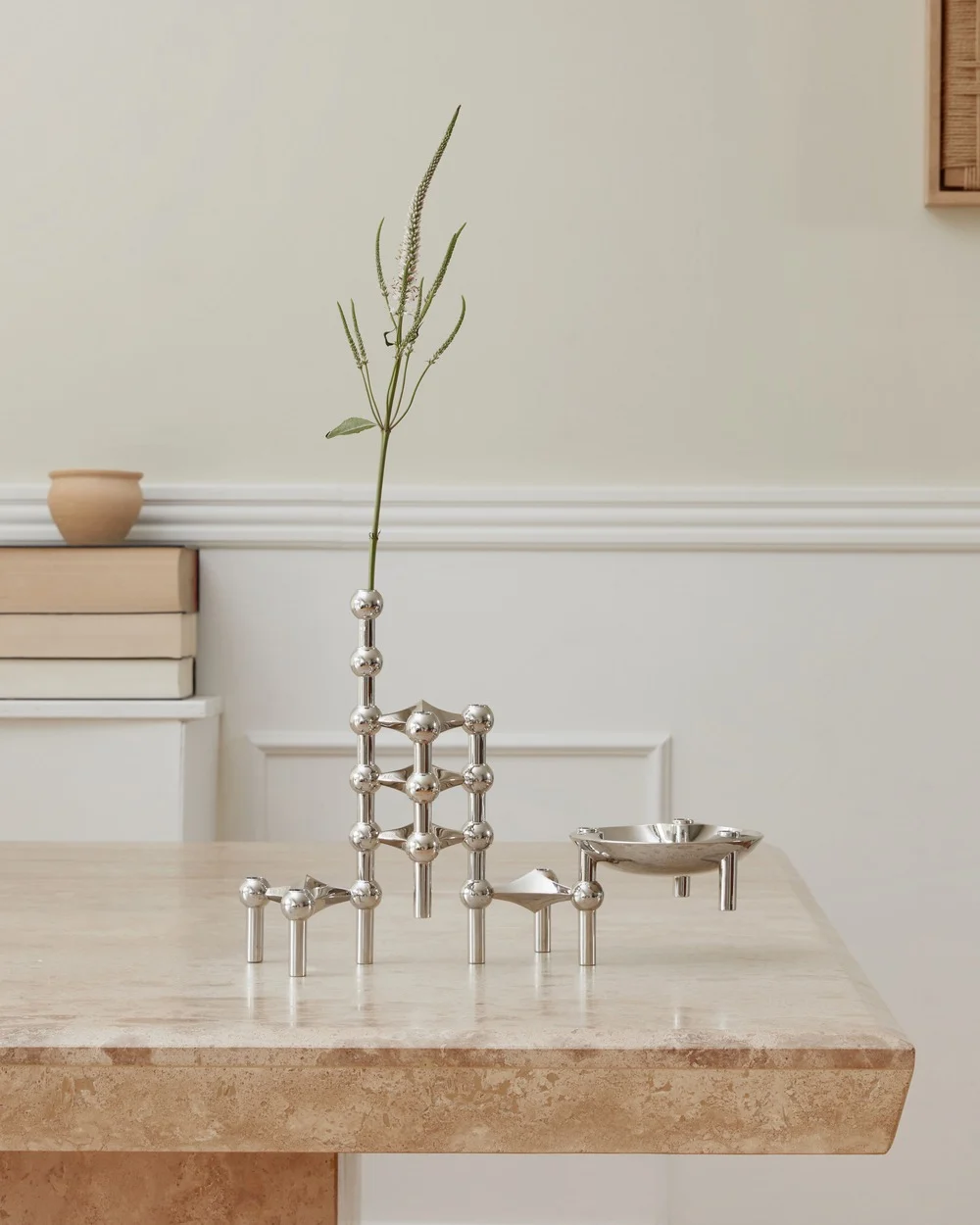 A polished silver modular candleholder system with a tall plant stem and a small, shallow dish, on a light brown stone table.
