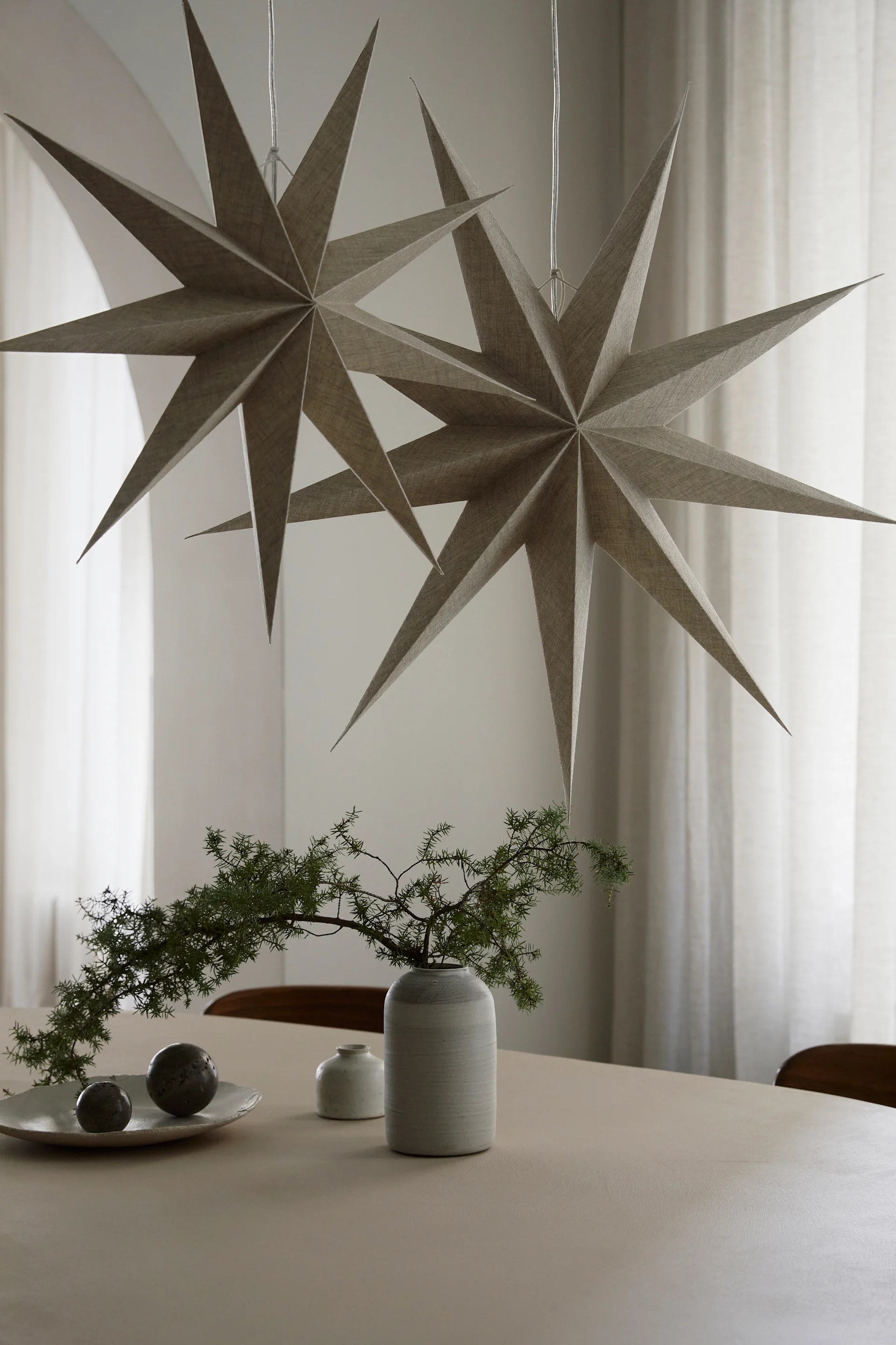 Two paper star lanterns from Watt & Veke with a natural linen finish hangover a dining table with a vase and some green plants. 