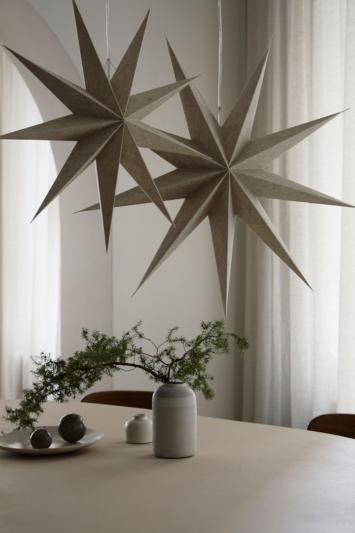 Two paper star lanterns from Watt & Veke with a natural linen finish hangover a dining table with a vase and some green plants. 