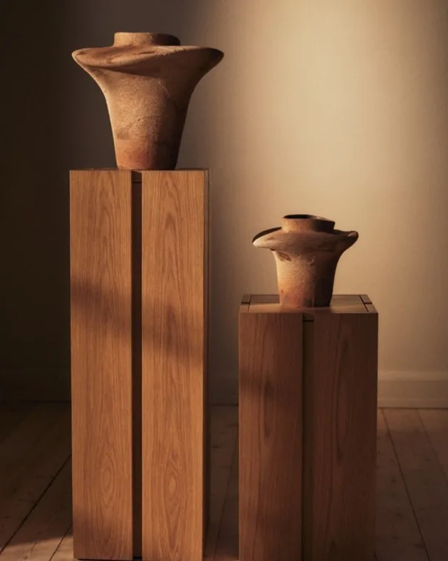 Two earthy, flared ceramic vases on light wooden pedestals.