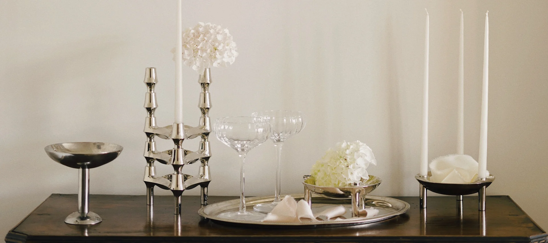 Elegant table display with silver candle holders, white candles, glass coupes, and white hydrangeas on a wooden surface.