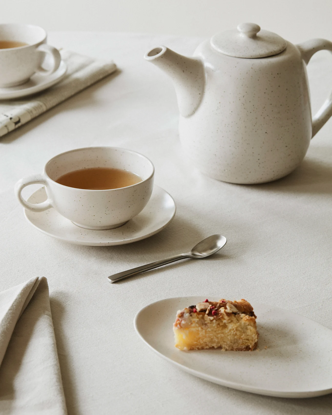 A minimalist scene with a white speckled teapot and teacup filled with tea, a spoon, and a slice of cake on a plate, all on a light linen tablecloth.