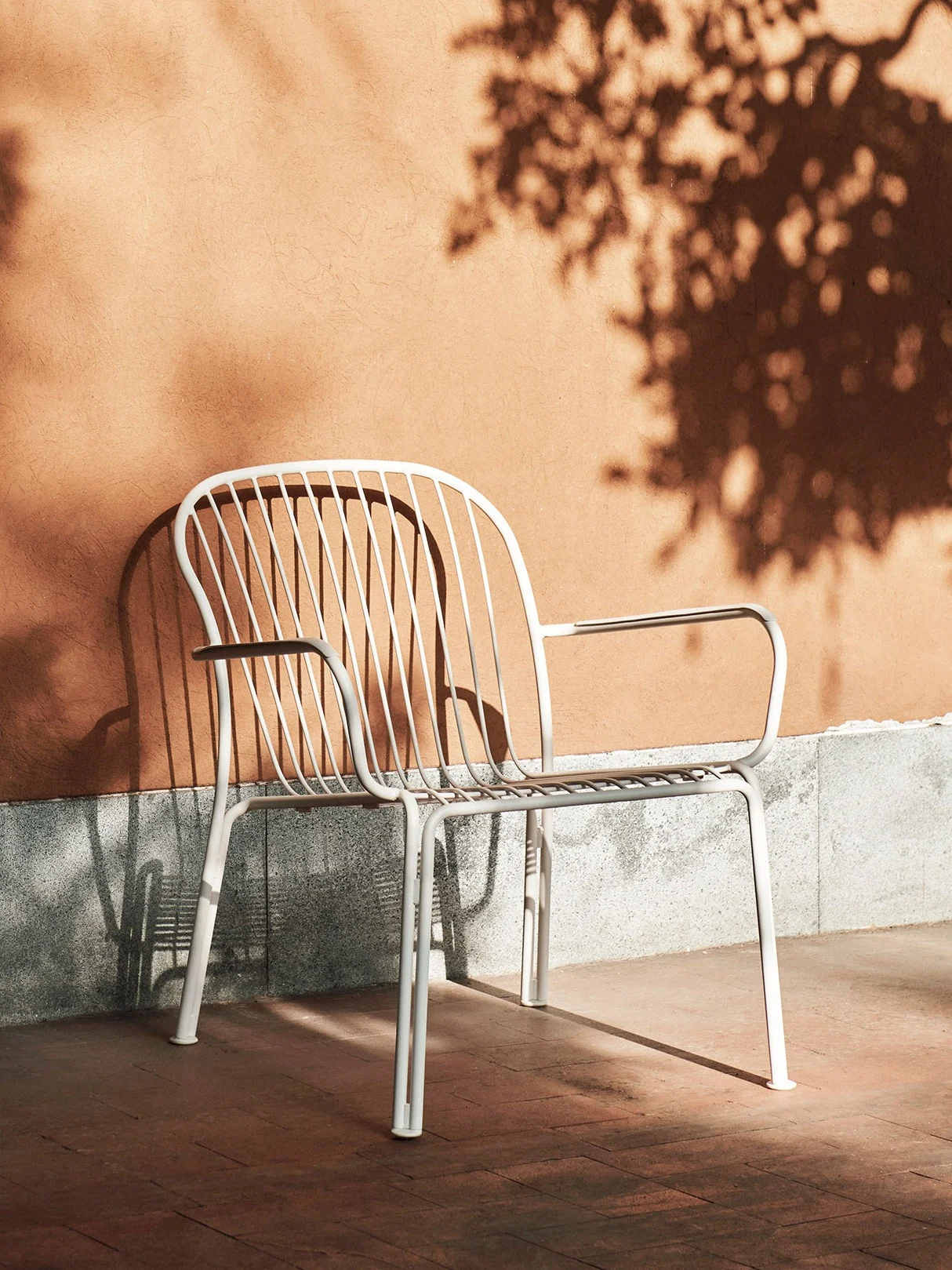White slatted metal outdoor chair against an orange wall with tree shadows on a sunny day.