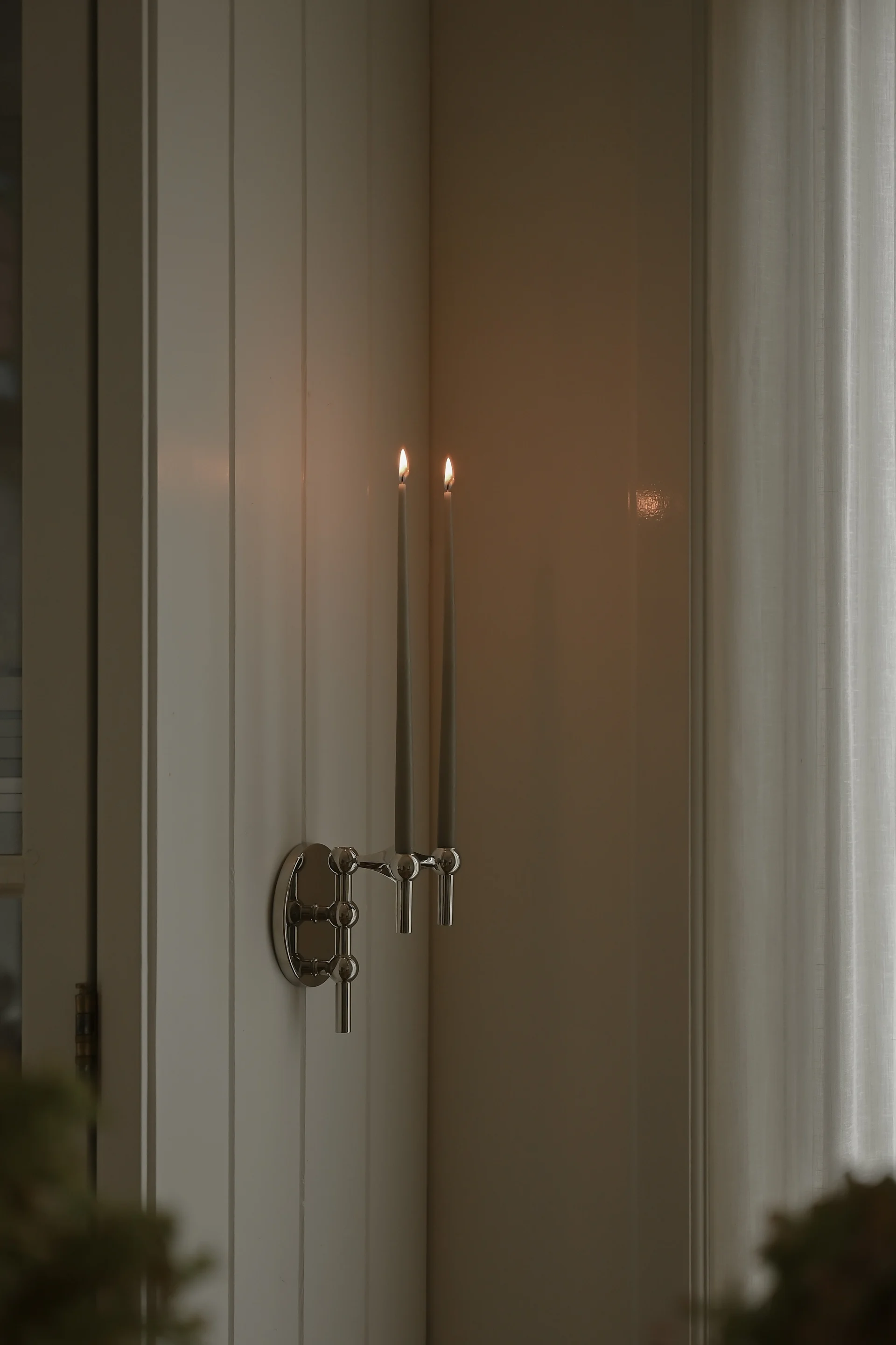 A silver wall-mounted candelabra holds two lit grey taper candles on a paneled wall next to a sheer curtain.