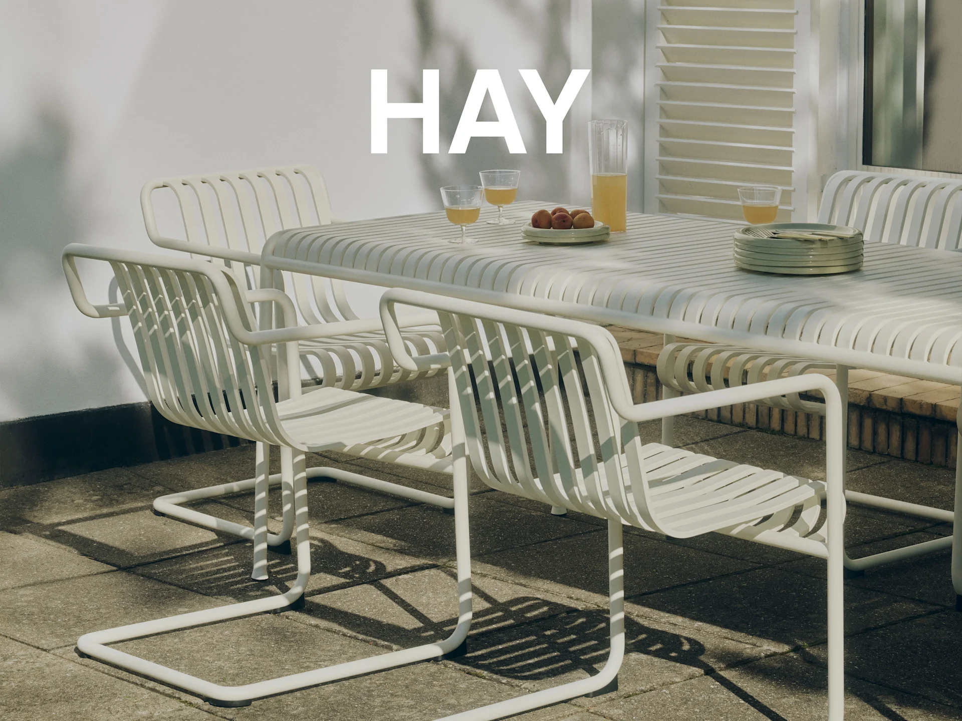 White slatted outdoor dining set on a sunny patio, with juice and apricots on the table. HAY logo is overlaid.