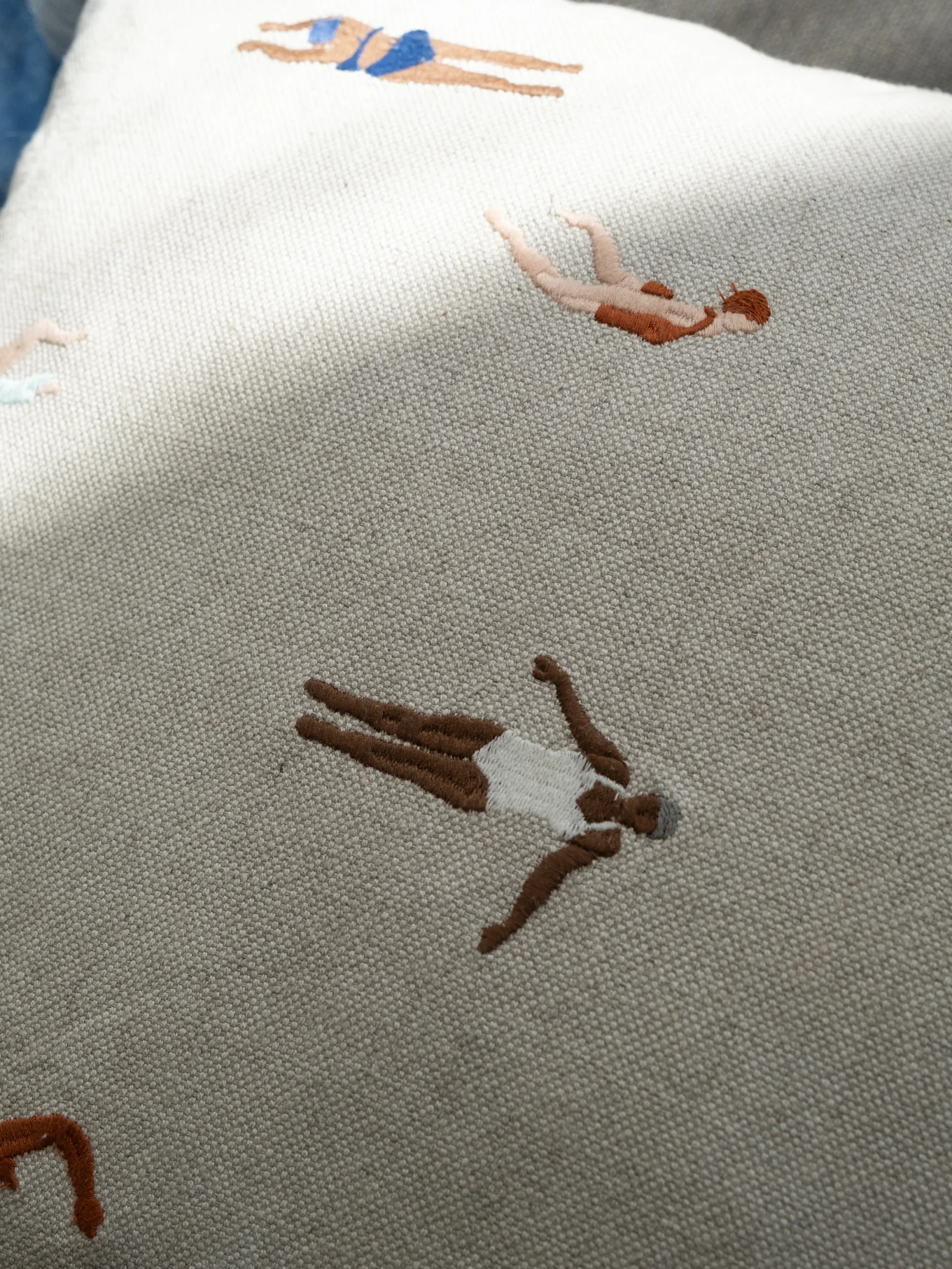 Light-colored embroidered fabric with stylized swimmers in diverse skin tones and swimwear.