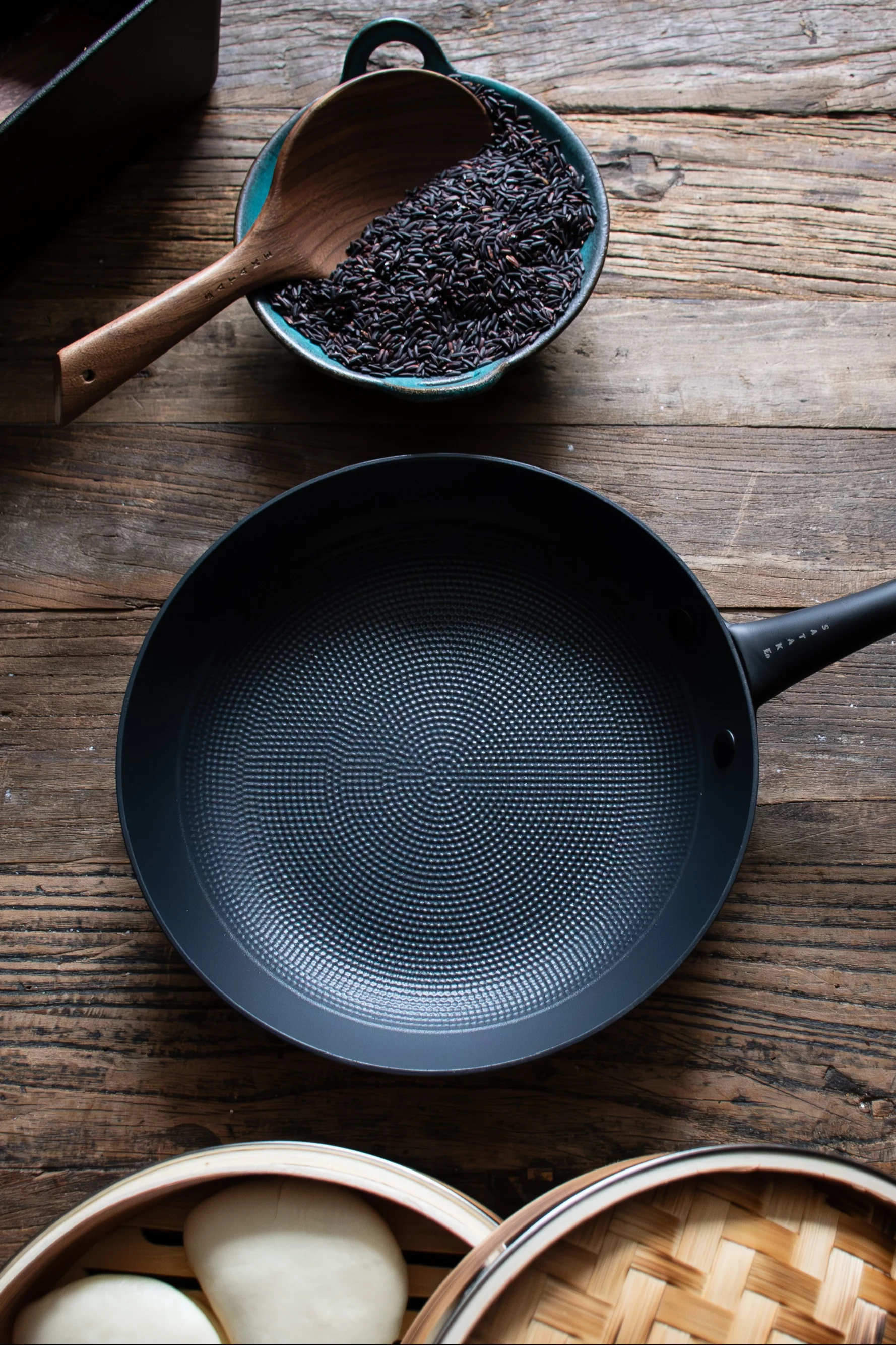 Overhead view of a black frying pan, a bowl of black rice with a wooden spoon, and a bamboo steamer with white buns on a wooden table.
