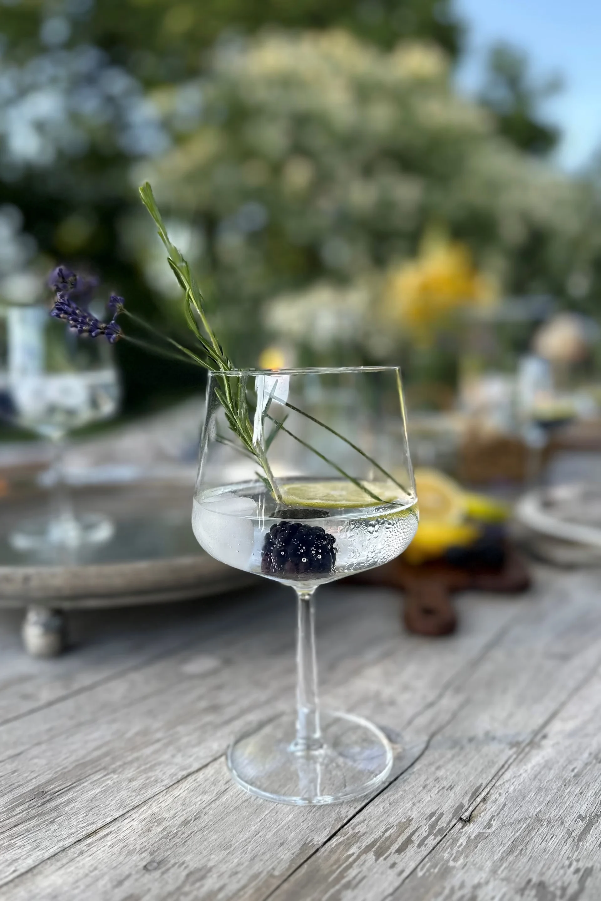 A stemmed glass cocktail with a blackberry, lime slice, lavender, and a green herb on a wooden table.