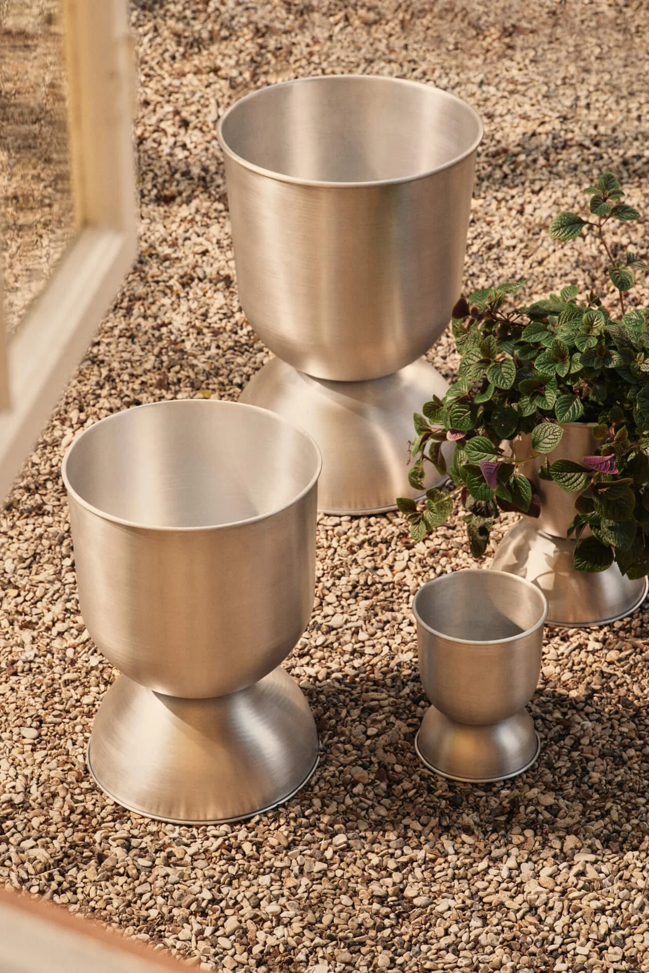Three brushed metal planters of different sizes on gravel, one containing a green plant.