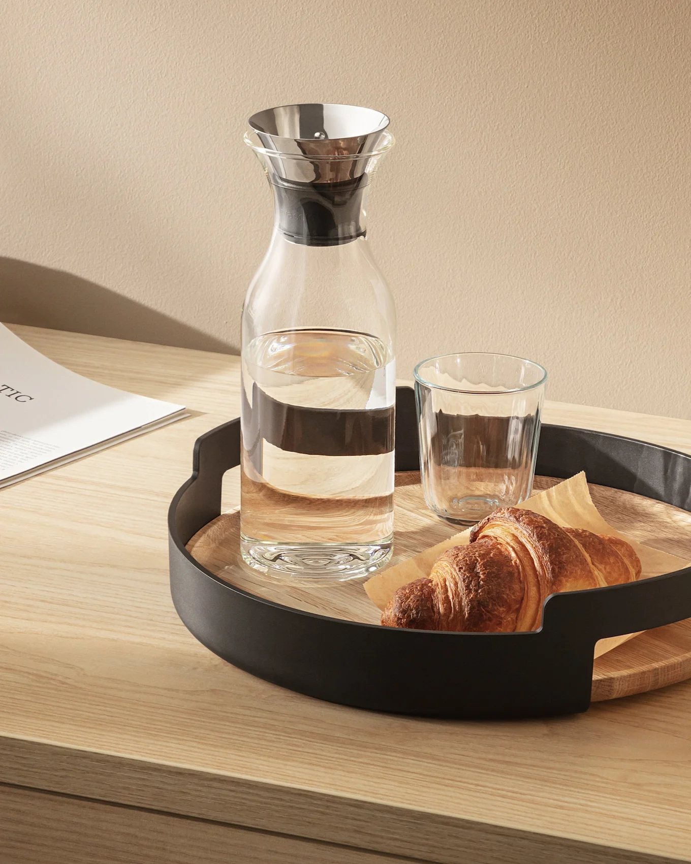 Glass carafe and water glass with a croissant on a round wooden tray with a black frame on a light wood table.