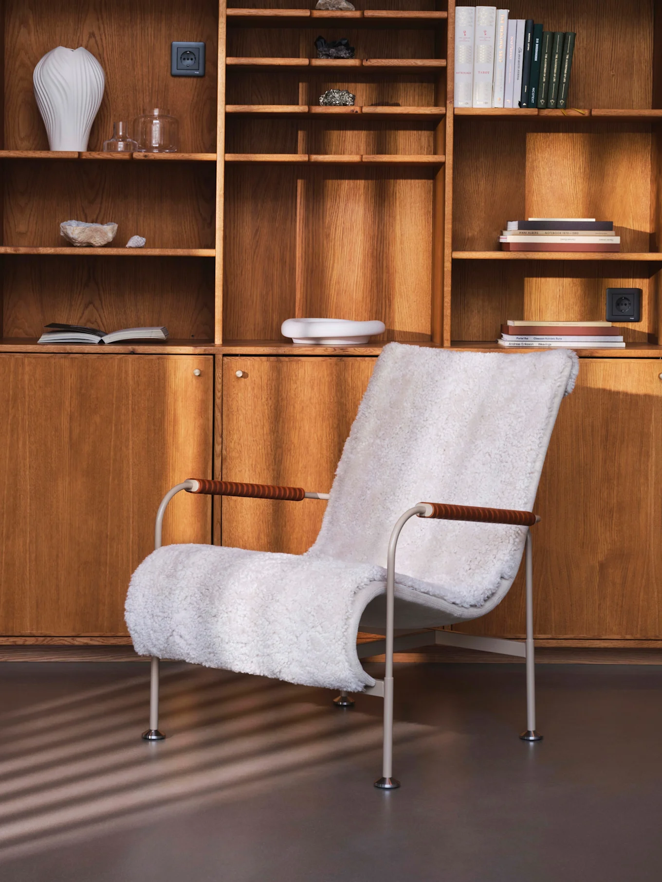 White faux-fur armchair in front of a wooden wall unit with shelves holding books, vases, and minerals.