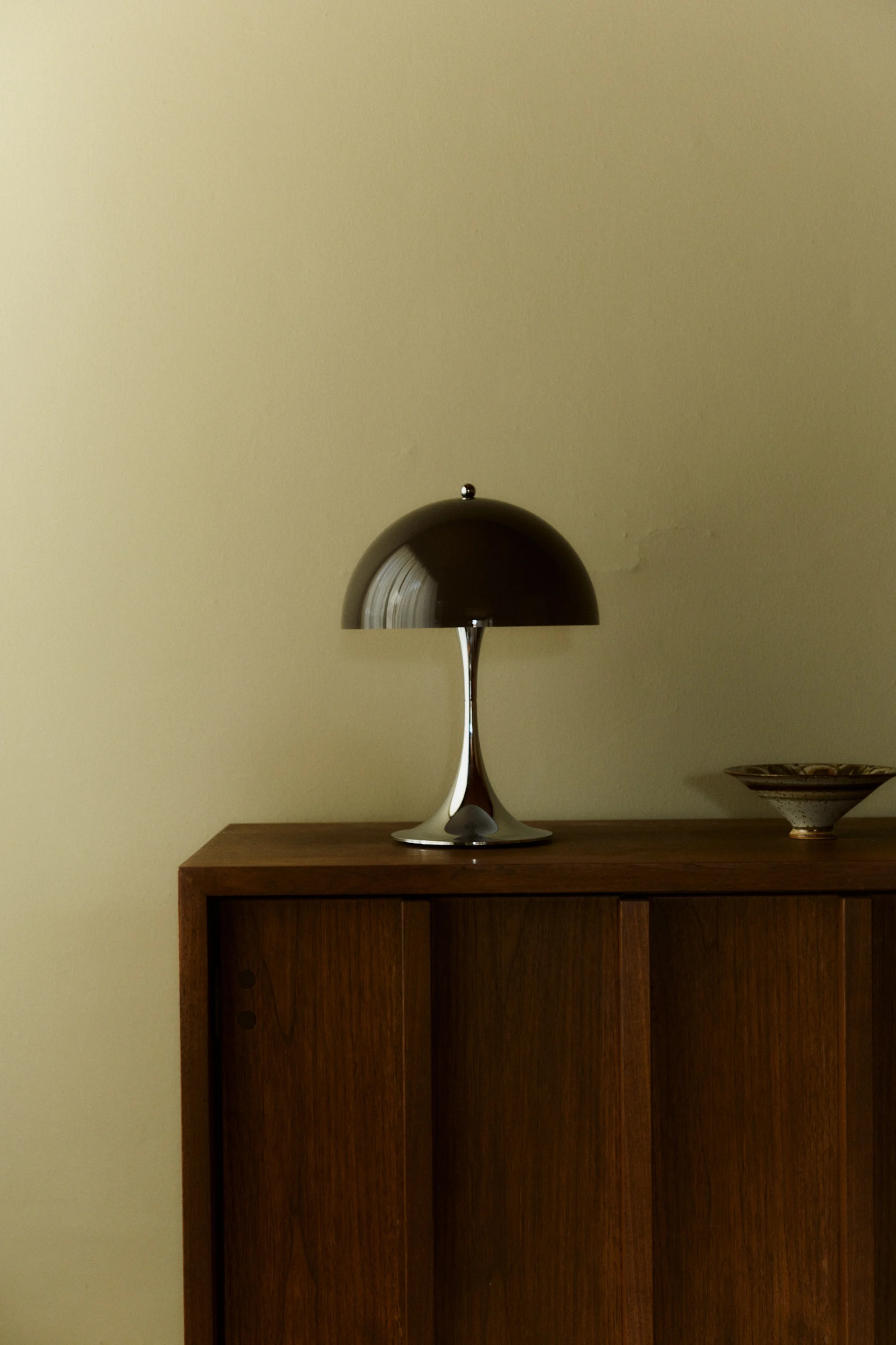 A sleek, modern mushroom table lamp with a dark dome shade and chrome base sits on a wooden cabinet next to a ceramic bowl.
