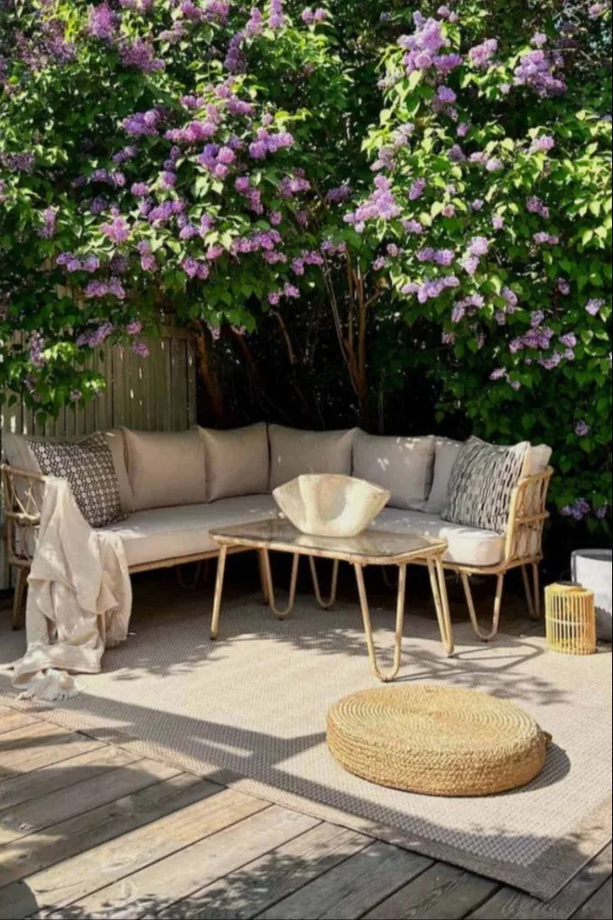 Outdoor patio with a woven corner sofa, coffee table, and pouf under a blooming lilac bush.