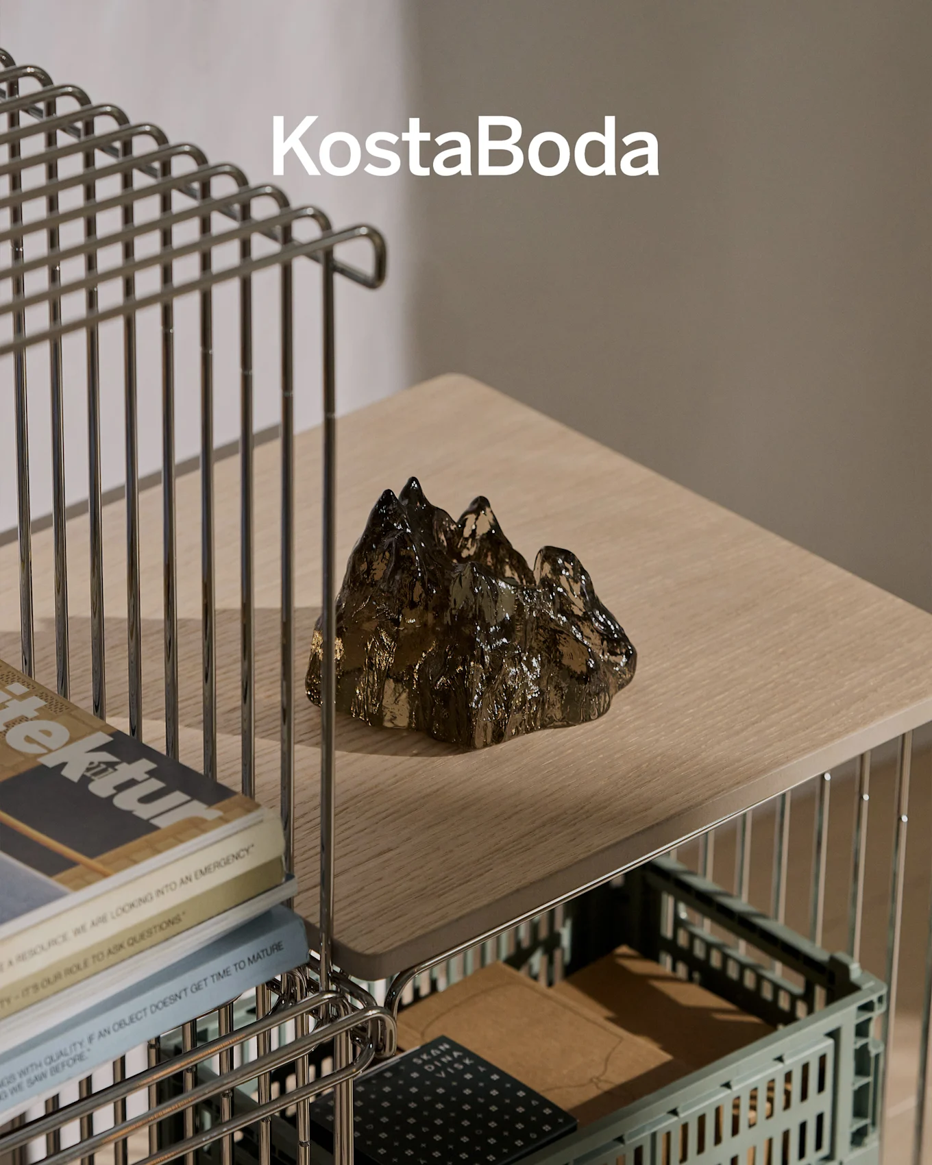 KostaBoda brand name above a dark, transparent, mountain-shaped glass sculpture on a light wooden shelf.